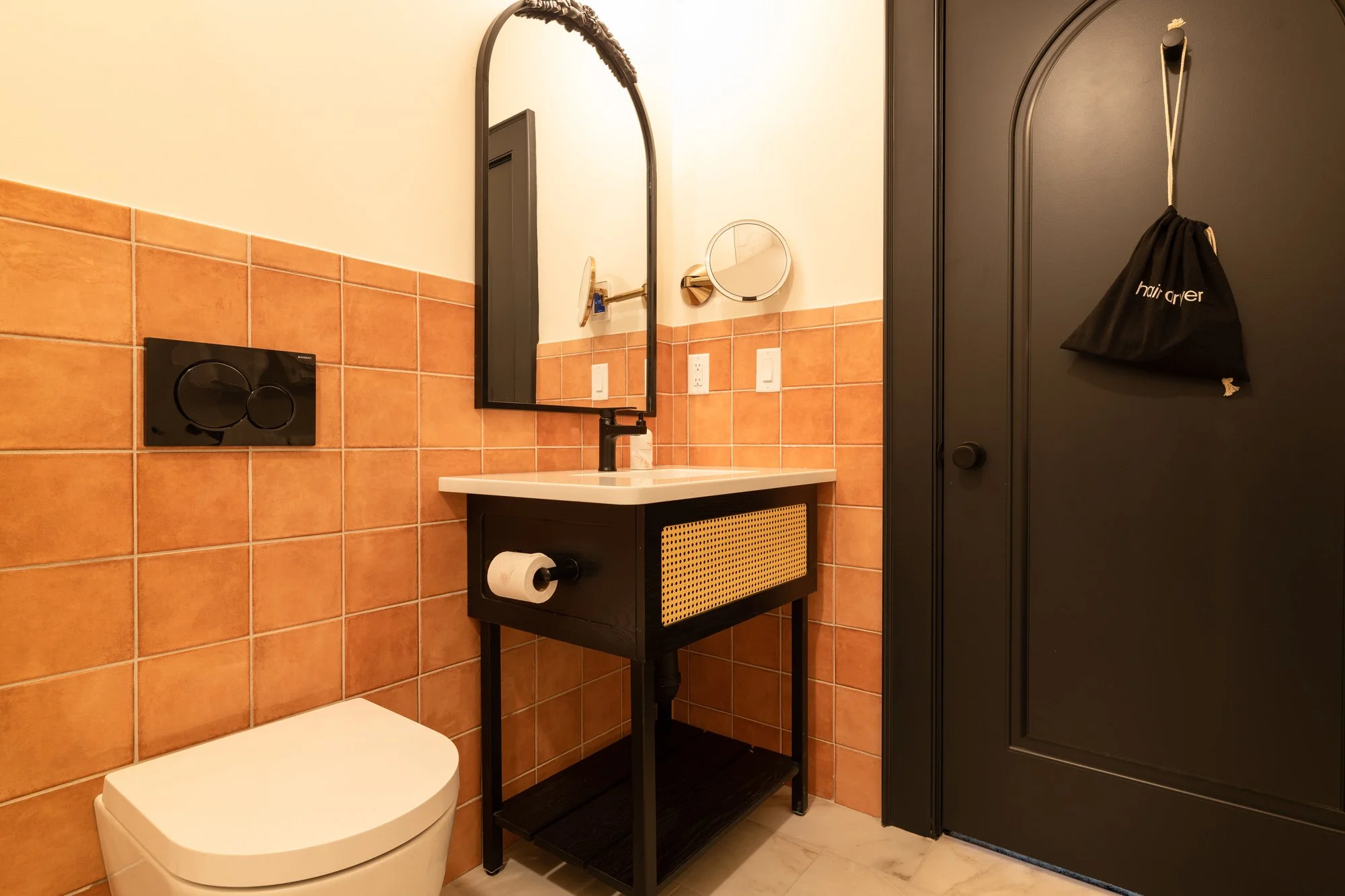 Small bathroom featuring a white toilet, a black and beige vanity with a sink and mirror, orange tiled walls, a black door with a hairdryer bag hanging on it, and a small round mirror on the wall.