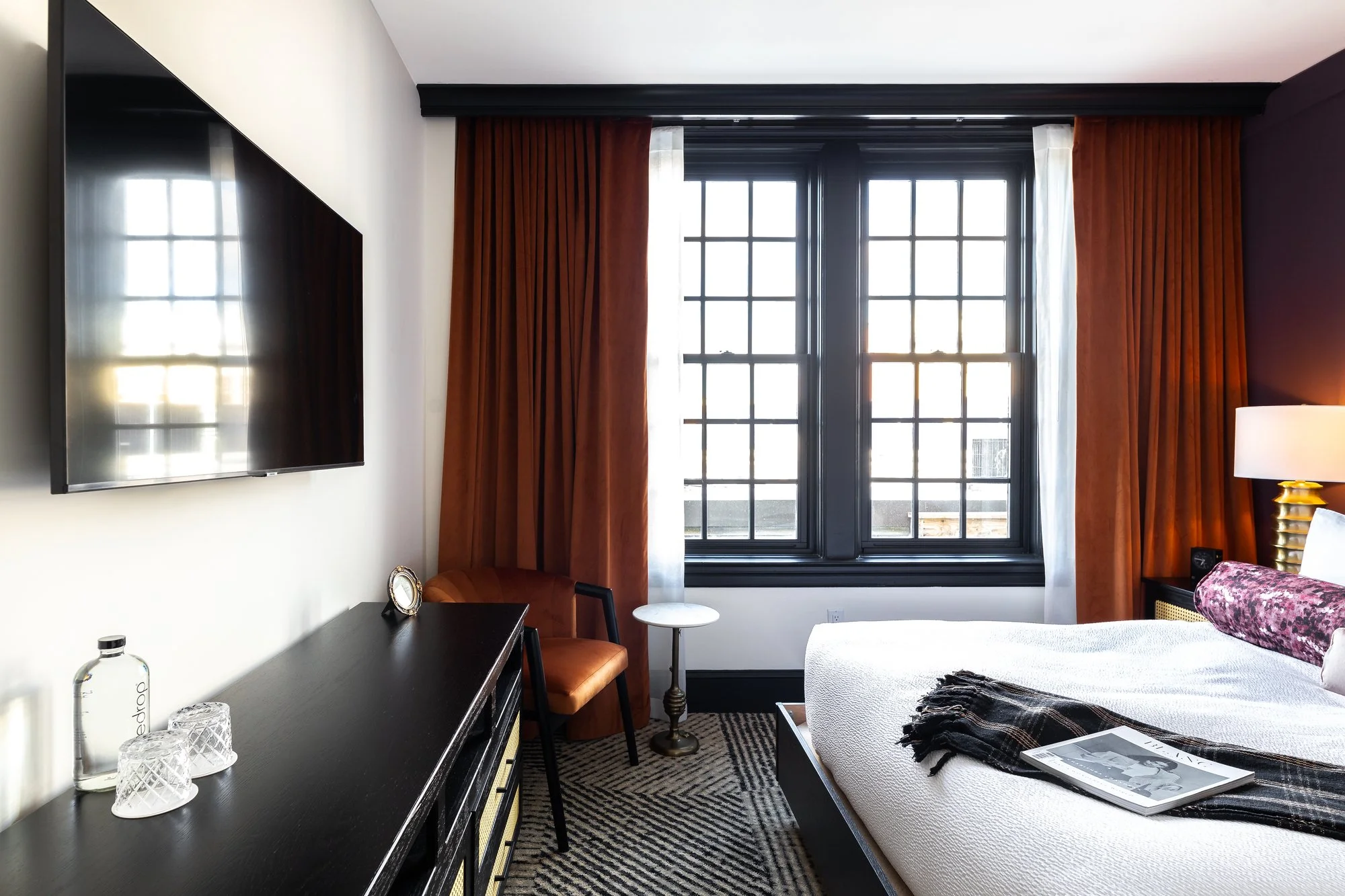 A modern hotel room with a large window, black window frames, rust-colored curtains, a flat-screen TV on the white wall, a dark wooden dresser with glassware and a water bottle, a small round side table, an orange chair, a bed with white bedding, a m