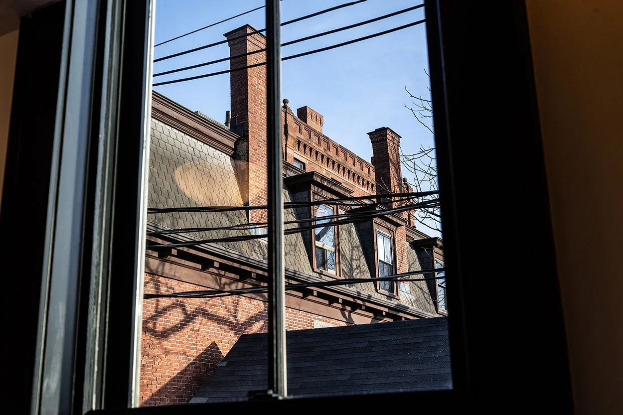 View of a brick building with chimneys and dormer windows seen through a window with black framing and blinds, clear blue sky in the background.