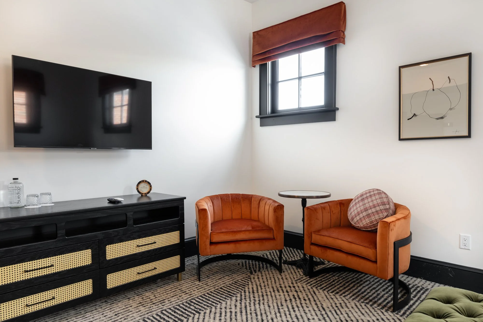 Living room with a wall-mounted flat-screen TV, a black and beige sideboard, two orange armchairs, a small round side table, a window with a red Roman shade, and artwork on the wall.