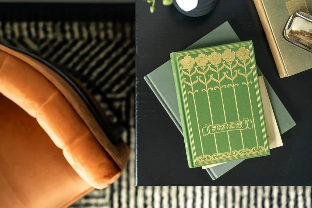 Close-up of a black table with a green book titled 'An Island Garden' by Cellia Thacher, other books underneath, a partially visible silver item, and a tan armrest of a chair or sofa, with a patterned black-and-white rug on the floor.