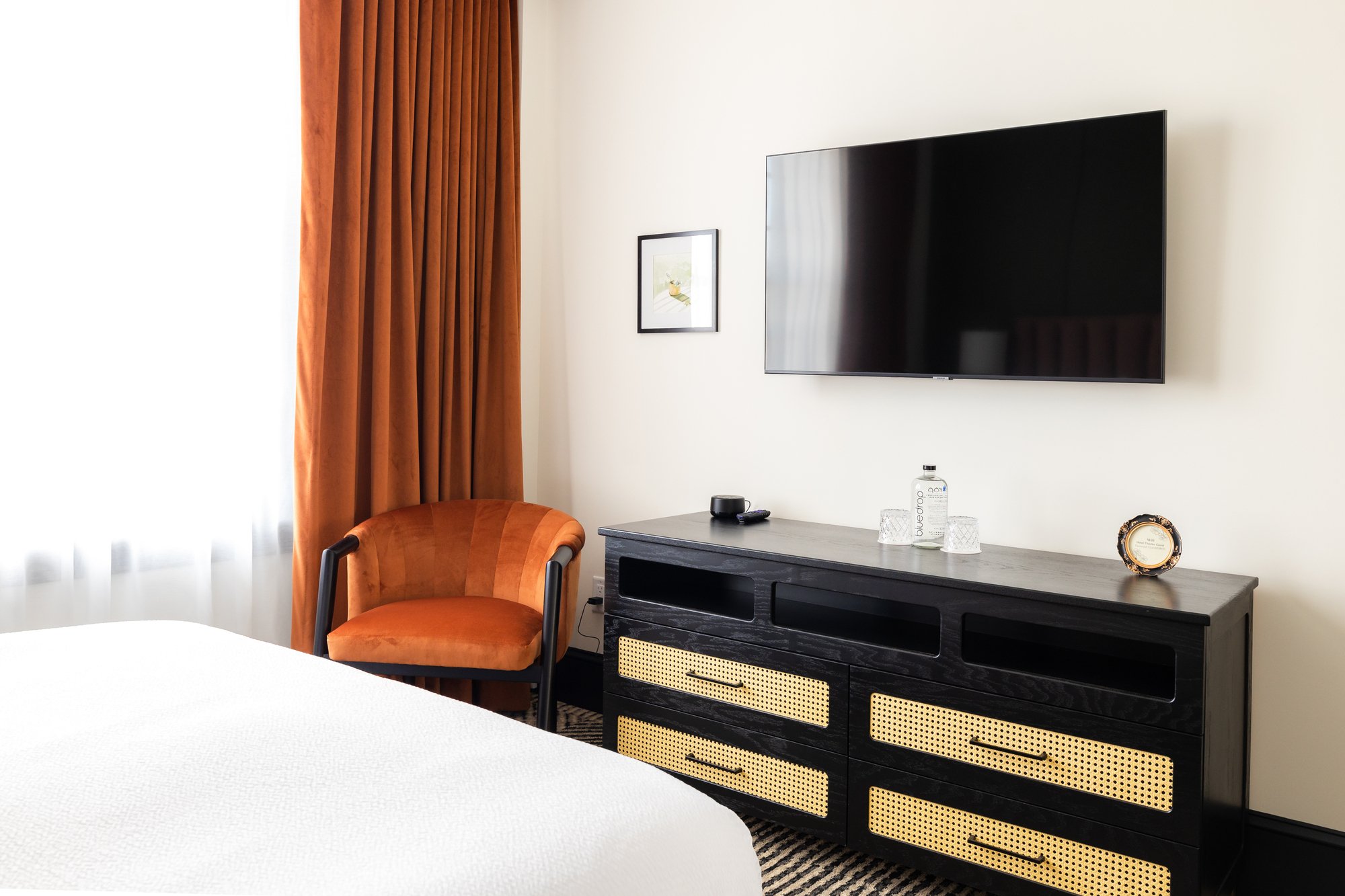 Hotel room with a flat-screen TV mounted on the wall, a black and tan dresser, a small framed picture, an orange upholstered armchair, and burnt orange curtains next to a window with sheer white curtains.