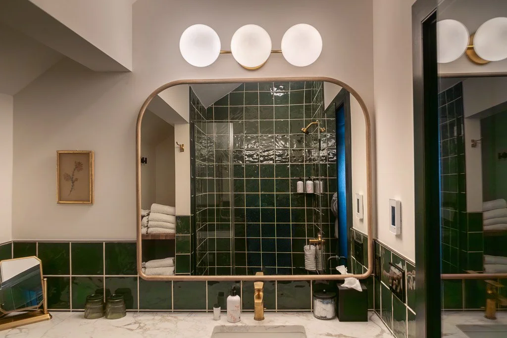 Bathroom vanity with dark green tile, gold framed mirror, and tiled shower in the Downtown Luxury Condo at Hotel Thaxter in Portsmouth, New Hampshire.