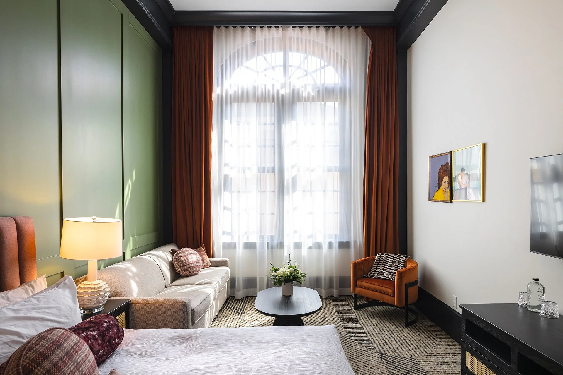 A hotel room with a large window, cream sofa, brown and plaid pillows, a side table with a lamp, an orange armchair, a black coffee table with a flower arrangement, a TV, and framed artwork on the wall.