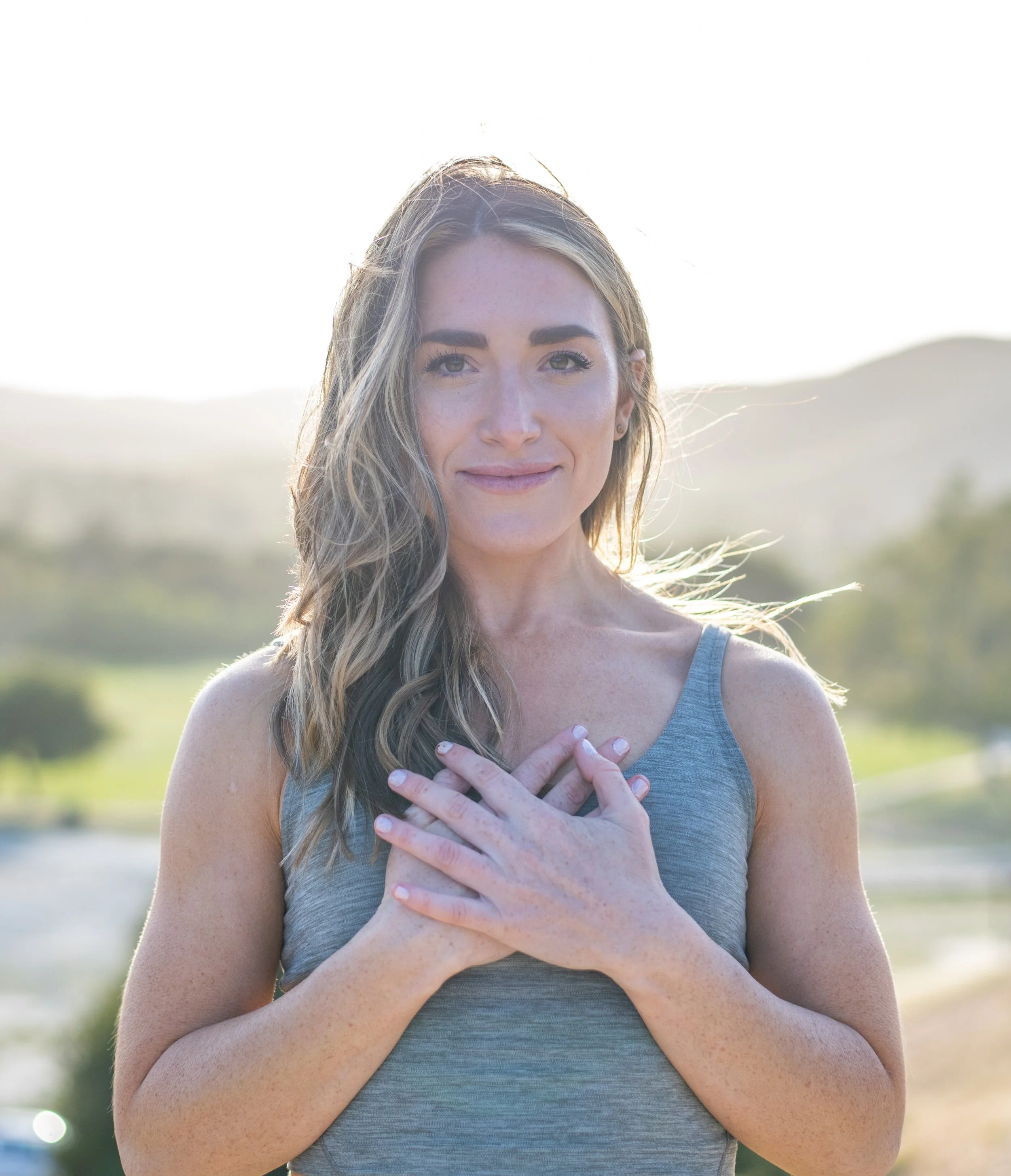 A woman with long, wavy hair smiling outdoors in a natural landscape, wearing a sleeveless gray top and holding her hands over her chest.