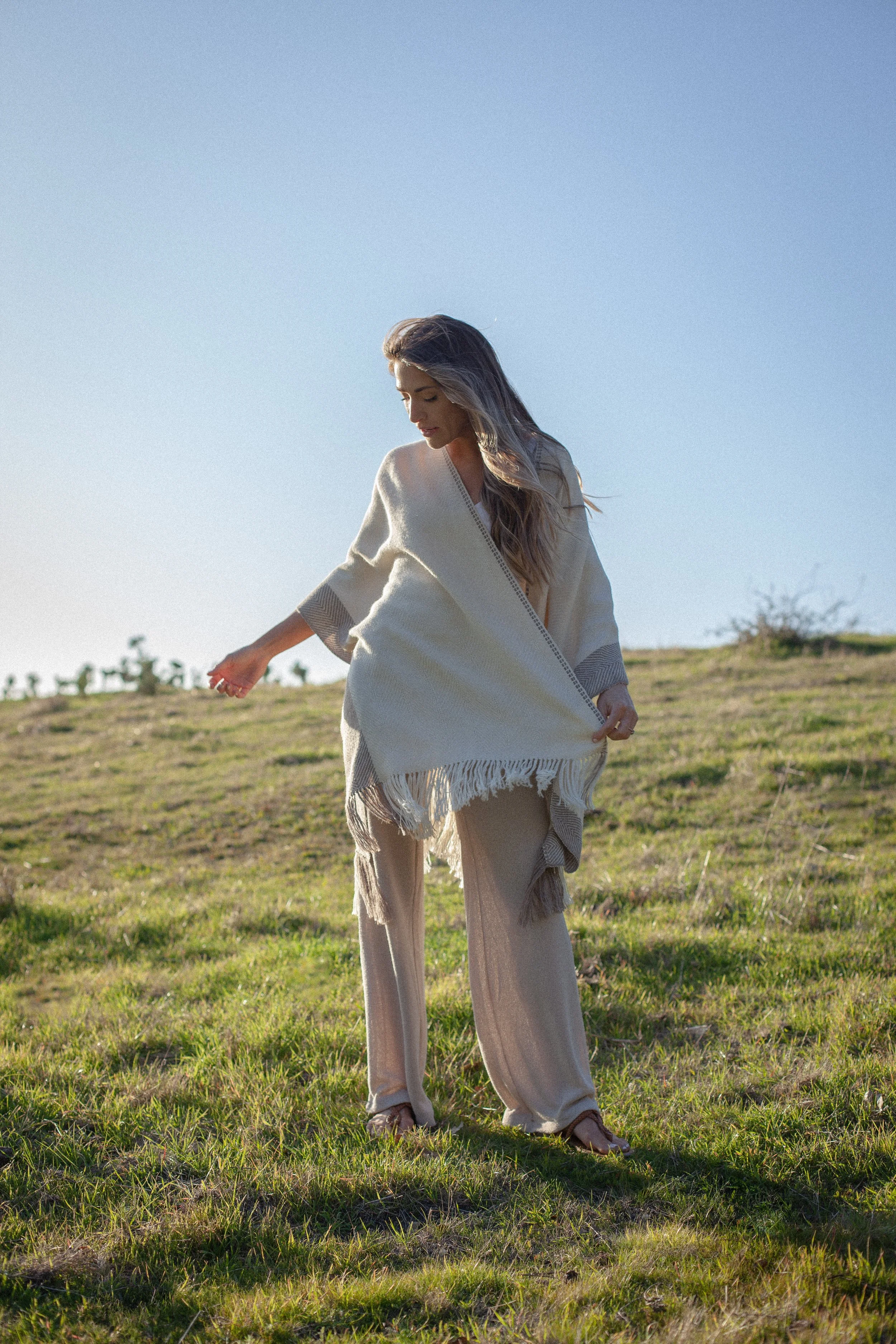 A woman standing outdoors in a grassy field during daylight, wearing beige wide-leg pants, a cream-colored shawl, and tan sandals, with her head slightly bowed and her hair flowing over her shoulders.