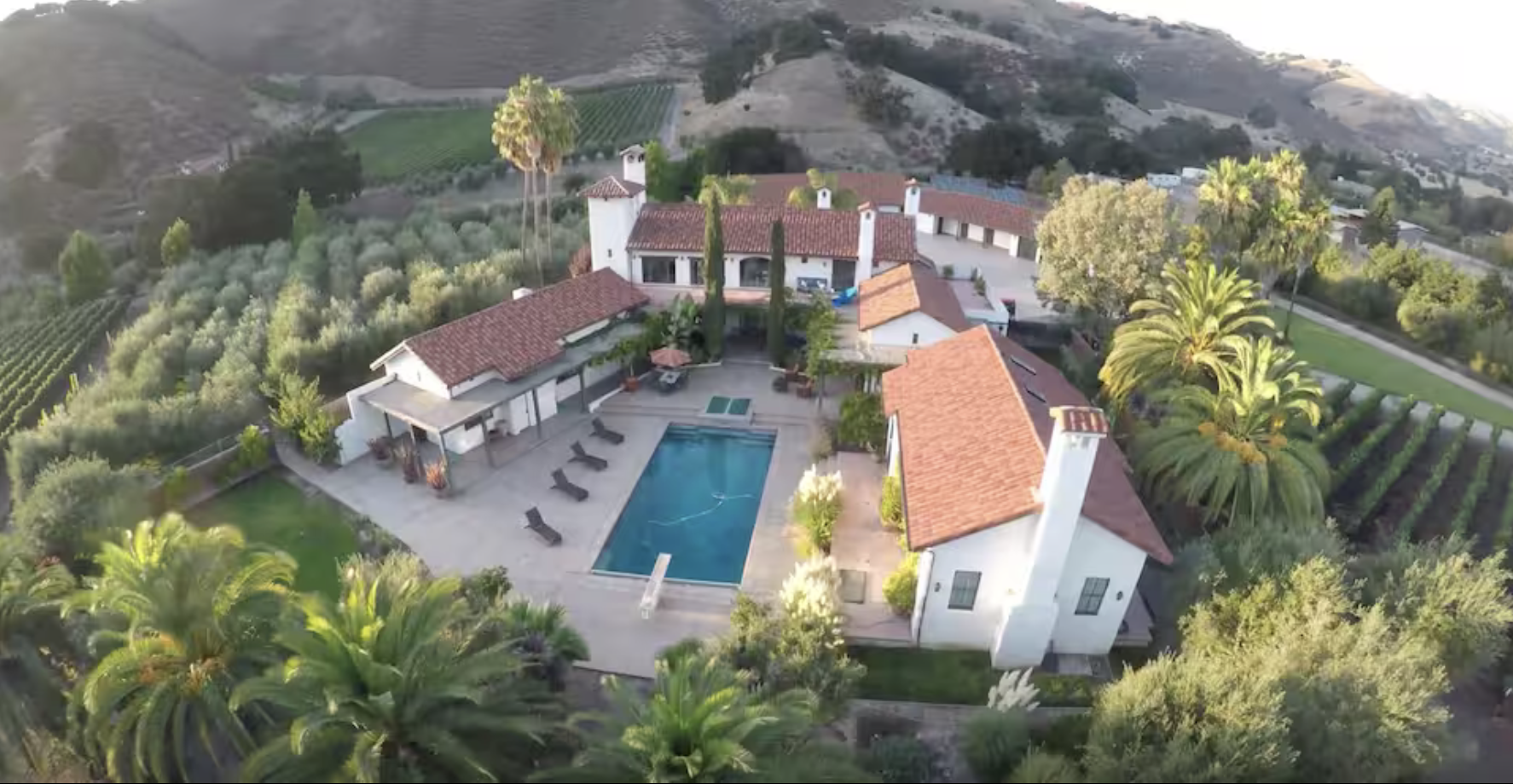 Aerial view of a luxurious Mediterranean-style estate with white stucco walls, red tile roofs, and a large swimming pool surrounded by lounge chairs. The estate is set amidst lush green trees, palm trees, and rolling hills with vineyards and farmland in the background.