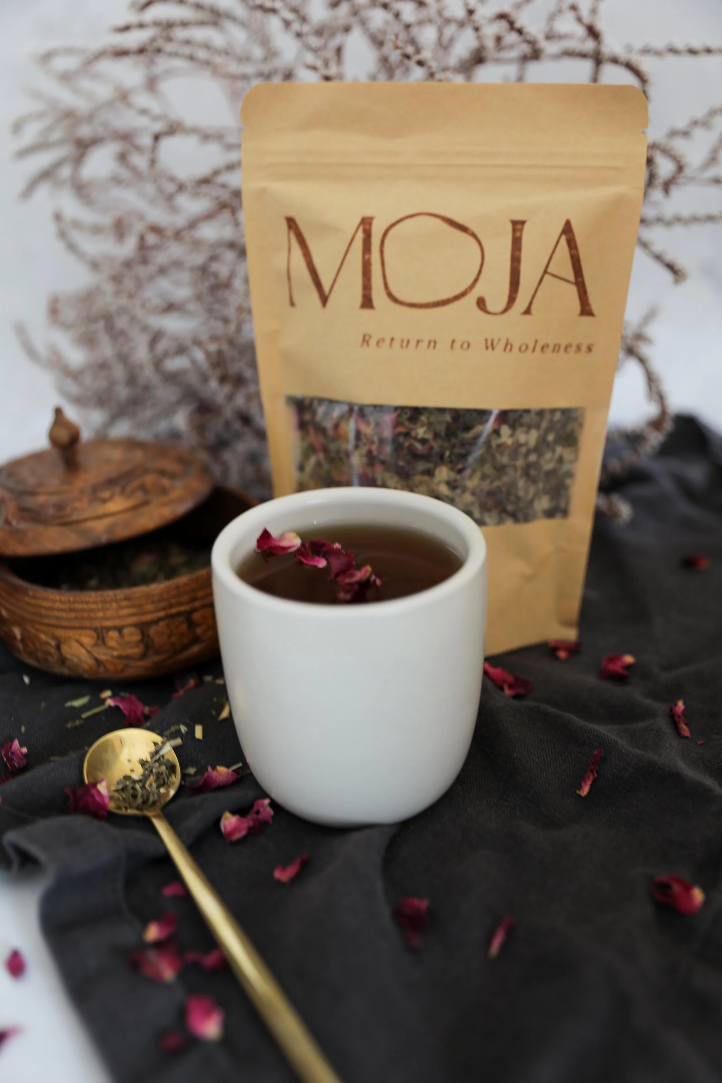 A white teacup filled with tea, garnished with rose petals. Behind it, a beige tea package labeled 'MOJA' with the tagline 'Return to Wholeness'. There is a wooden bowl filled with dried herbs or tea leaves and a gold spoon on a black cloth, with rose petals scattered around. A blurred, snow-covered tree branch is in the background.