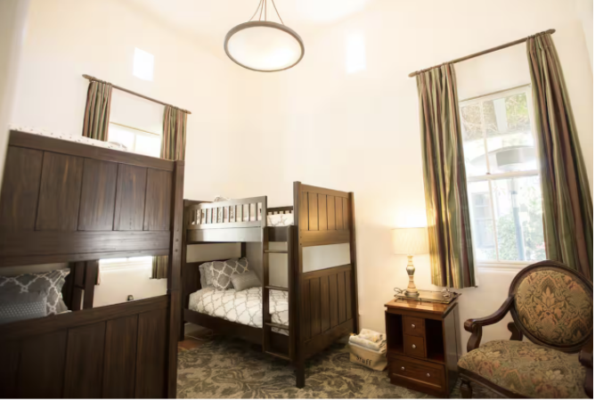 A bedroom with a bunk bed, a wooden chair with floral upholstery, a nightstand with a lamp, and a window with curtains.