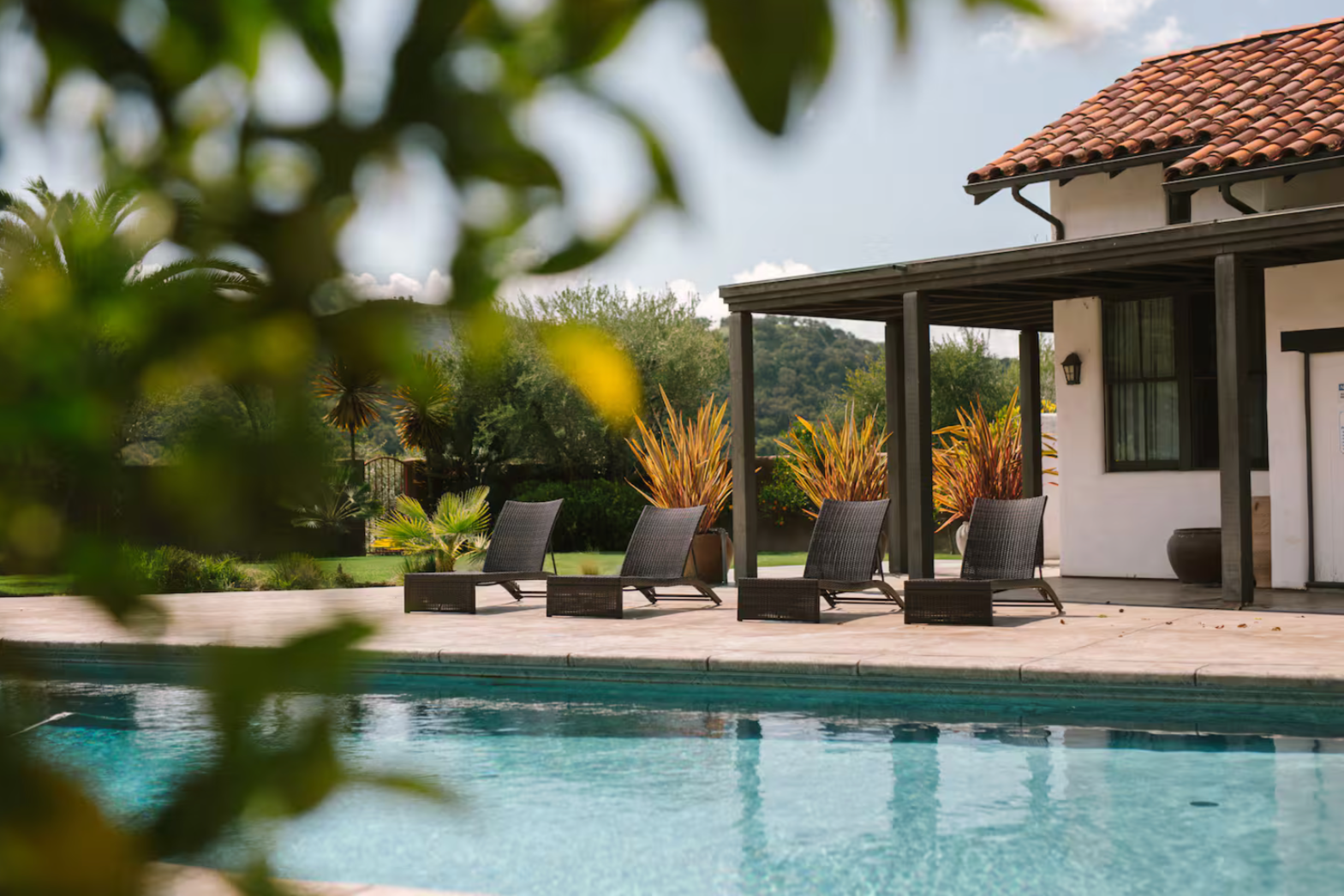 Poolside patio with four black lounge chairs, surrounded by plants and a house with a tiled roof and patio cover, in a sunny outdoor setting