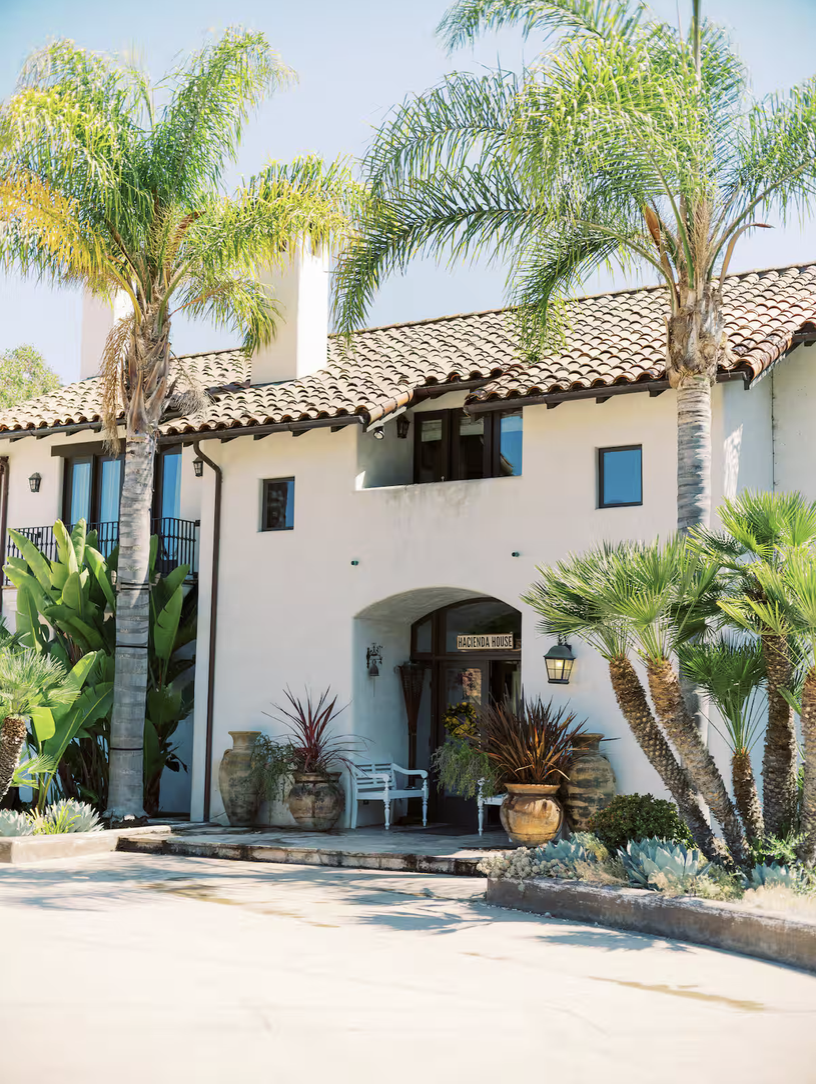 A white Mediterranean-style house with a tiled roof, surrounded by palm trees and desert plants, with a paved walkway leading to the entrance with potted plants and a sign that says 'Hacienda House'.
