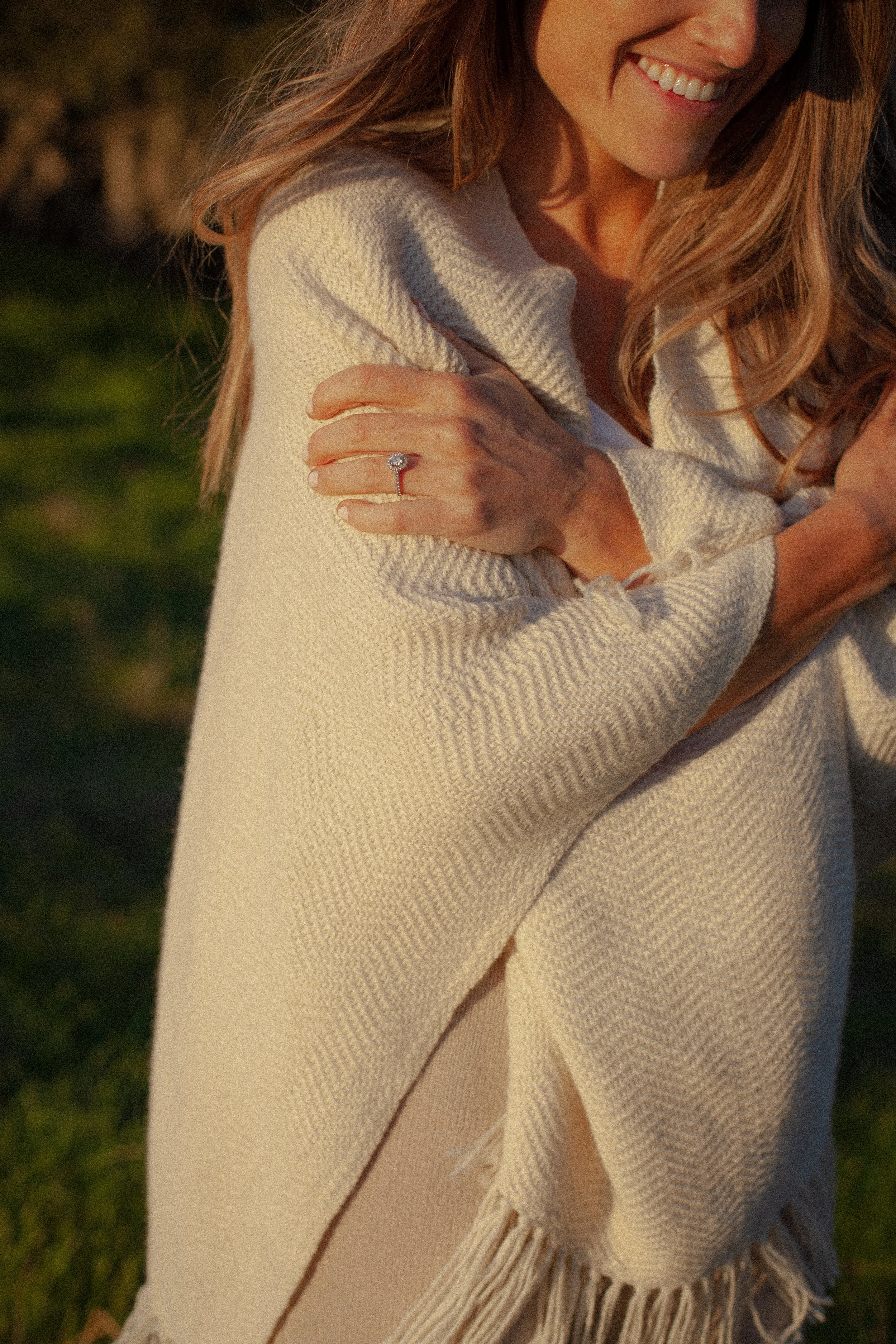 Close-up of a smiling woman with long, wavy, reddish-brown hair, wearing a black hat and a white knitted sweater with fringe. She is outdoors, holding her arm wrapped across her chest, showing an engagement ring with a large round diamond. The background is blurred greenery.
