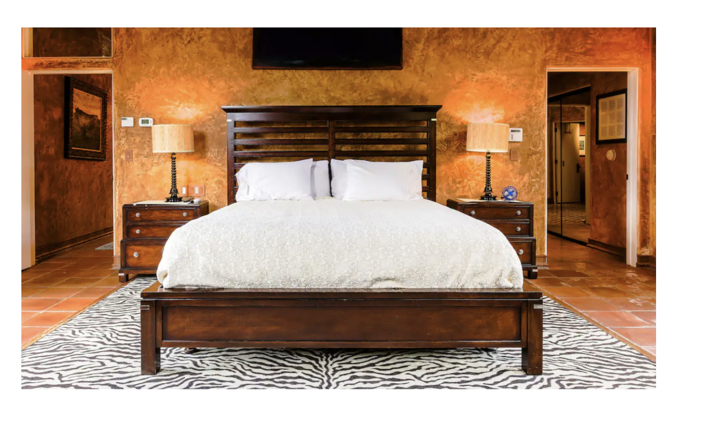 A bedroom with a large bed, white bedding, and a wooden headboard, flanked by matching nightstands with lamps, in a room with orange textured walls and a zebra-patterned rug.