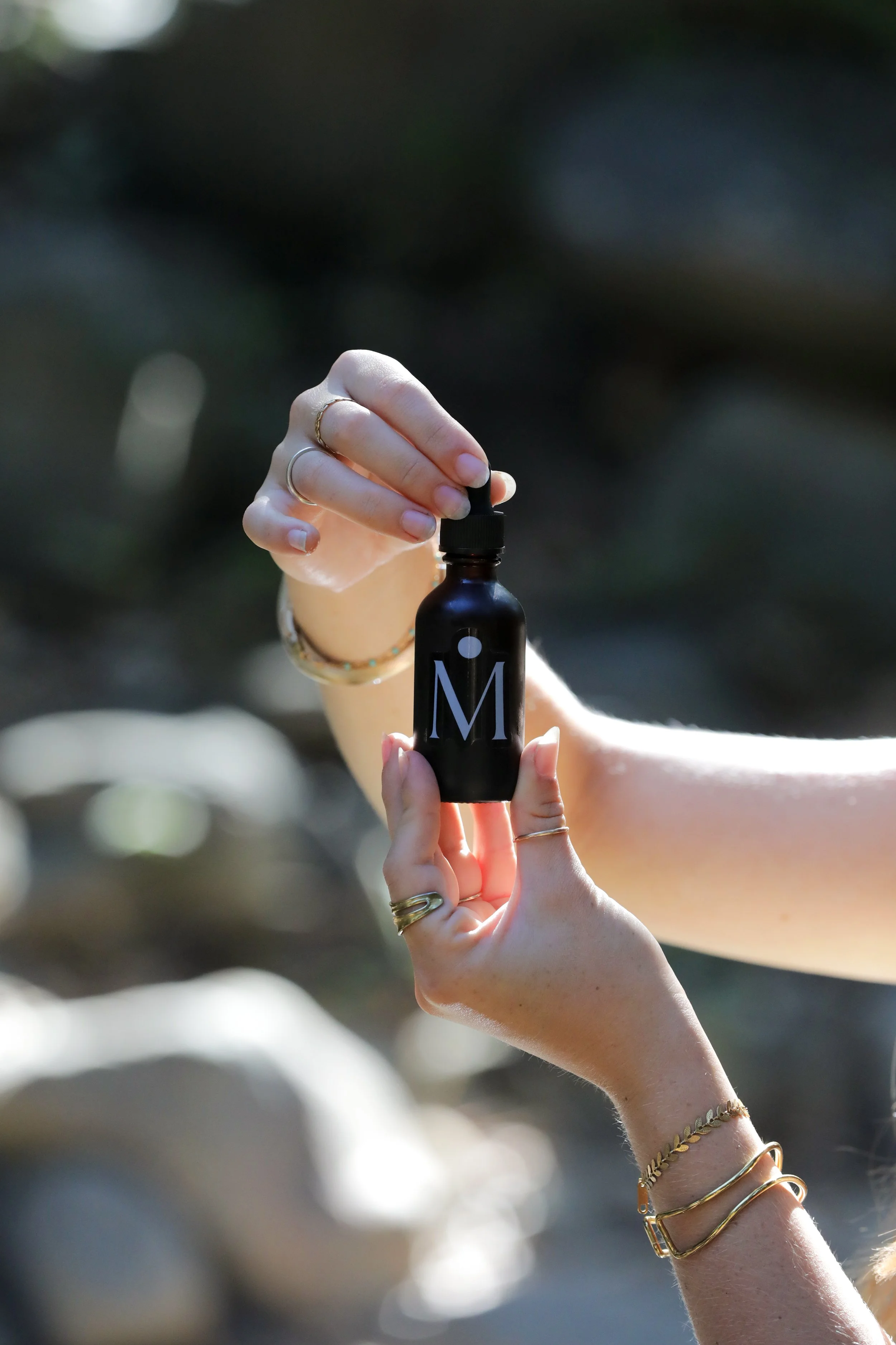 Person holding a small black bottle with a dropper cap, labeled with a large letter 'M', outdoors with blurred background.