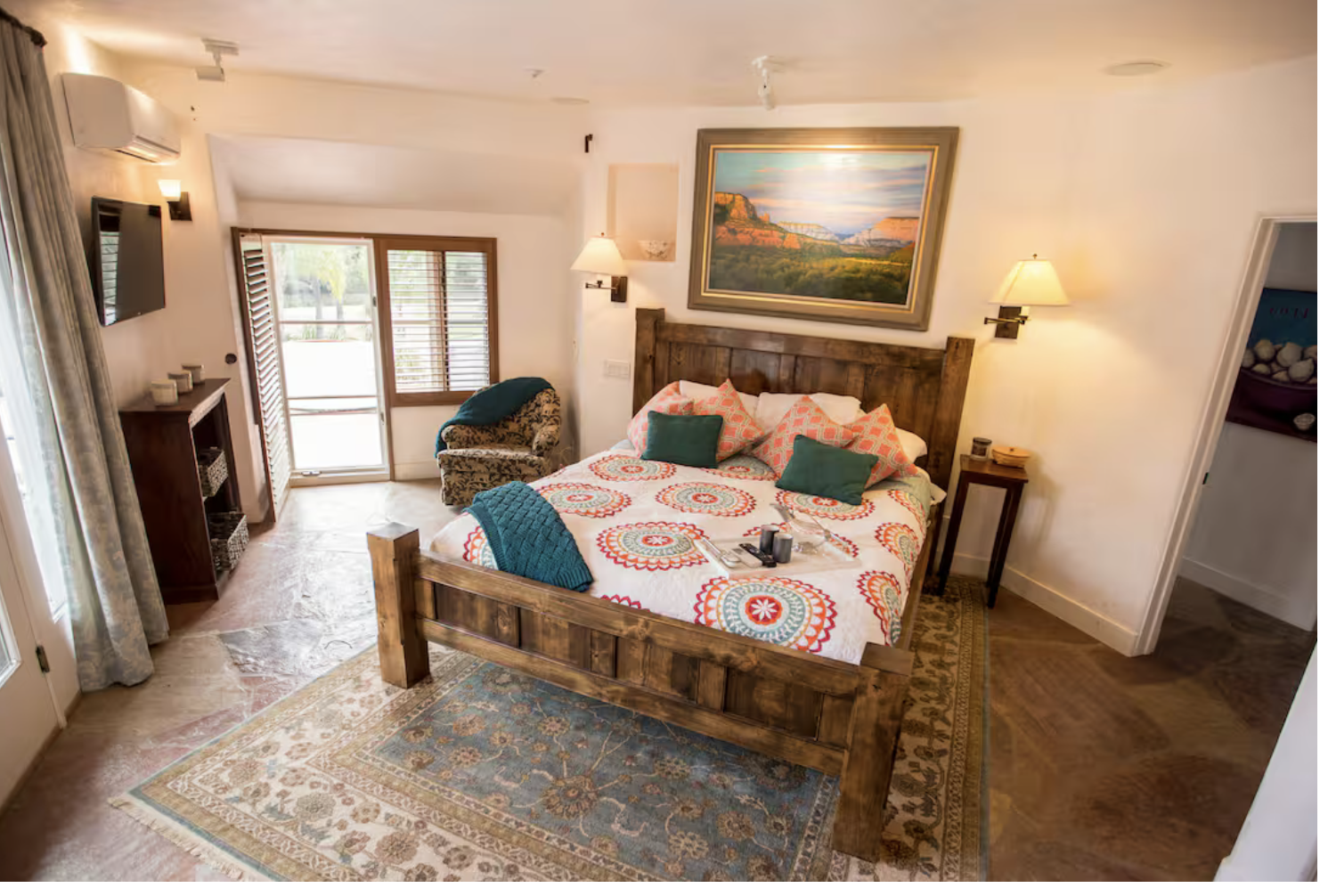 A cozy hotel bedroom with a wooden bed, colorful patterned bedding, and pillows. There is a patterned armchair near a sliding glass door with wooden shutters, and a landscape painting above the bed. The room has side tables with lamps, a small rug, and a flat-screen TV on the wall.