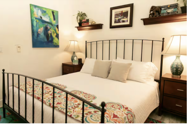 A bedroom with a black metal bed frame, white bedding, and three neutral-colored pillows. Two wooden nightstands with lamps are on each side of the bed. There are two wall-mounted shelves with decorative items and framed artwork on the wall above the bed. A colorful abstract painting is on the left wall.