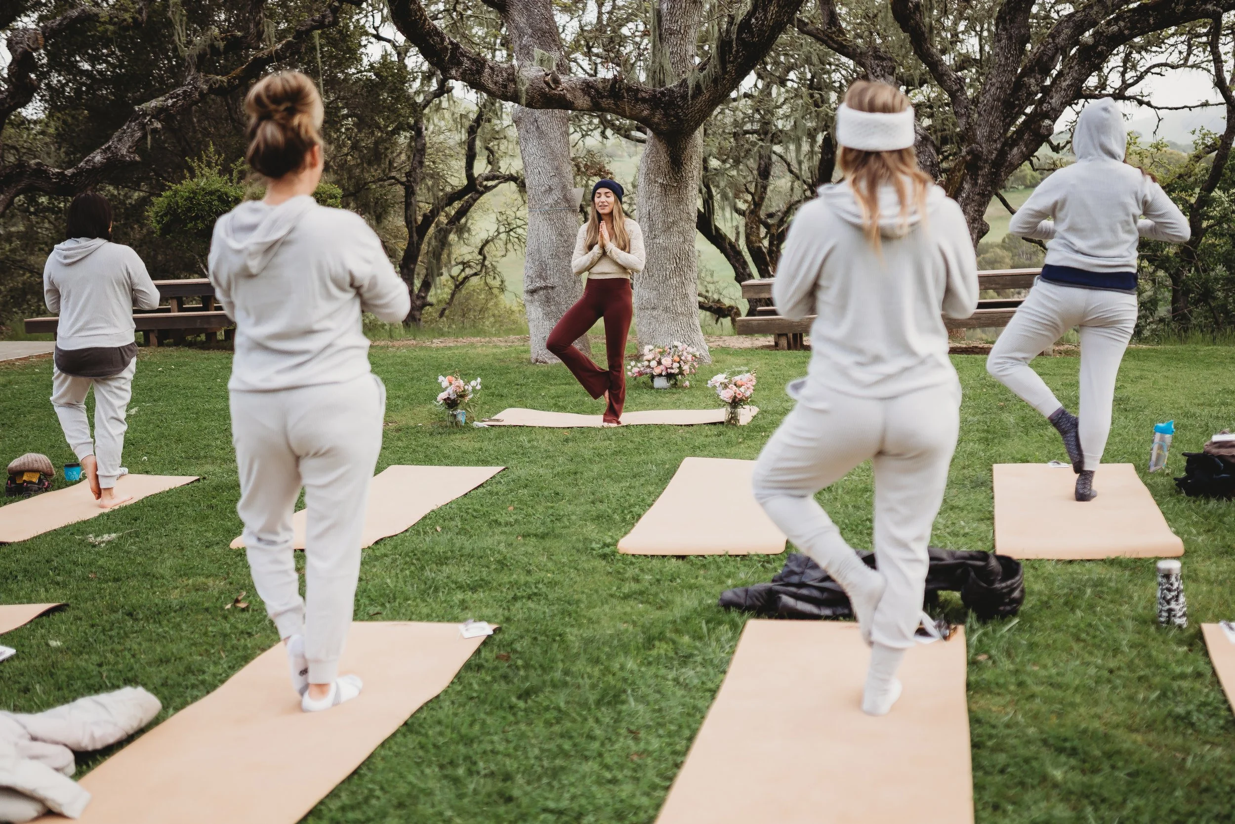 A group of people practicing yoga outdoors on yoga mats in a park with large trees, led by an instructor standing in front, all dressed in comfortable clothing.