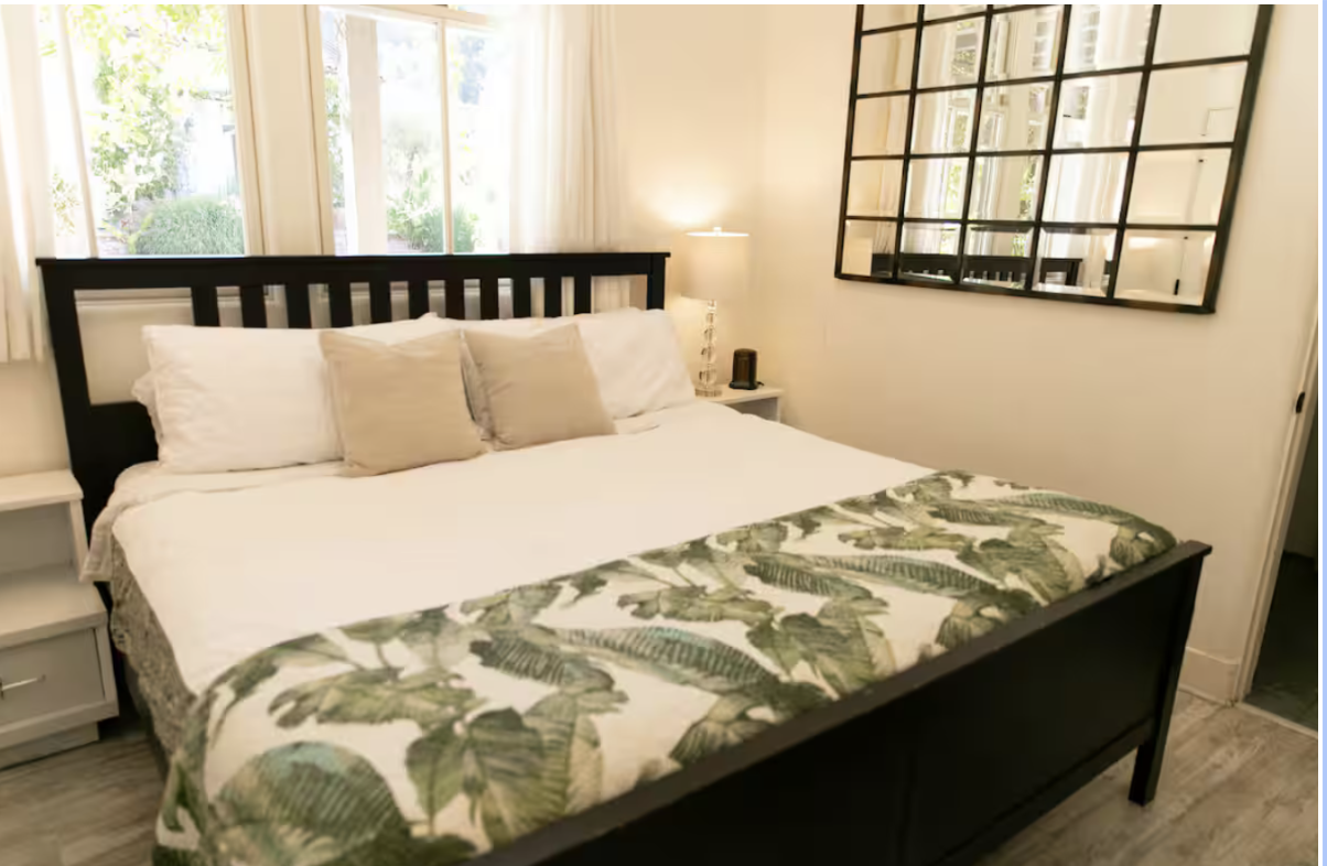 A cozy bedroom with a black wooden bed frame, white bedding, and decorative pillows. There is a patterned throw with green leaves at the foot of the bed. A nightstand with a lamp and small clock is beside the bed. Large windows behind the bed let in natural light, and a large wall mirror is on the right side.