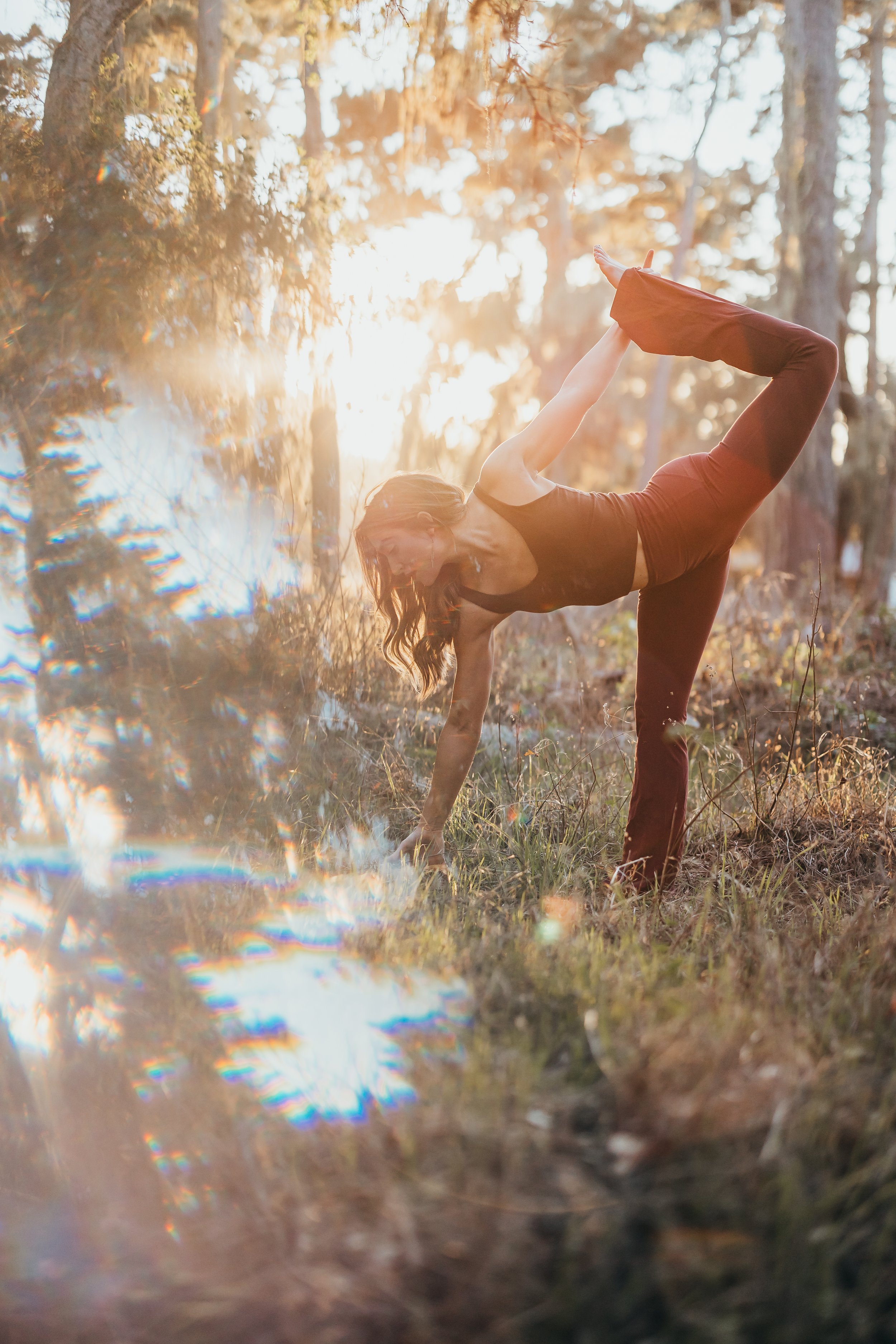 A woman practicing yoga outdoors in a wooded area during sunset, balancing on one leg with her other leg raised behind her, and her hand reaching for her foot.