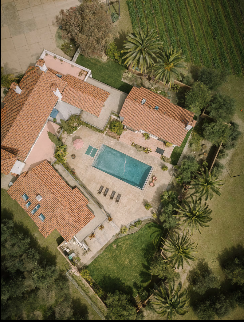 An aerial view of a backyard with a swimming pool, surrounded by multiple single-story homes with red-tile roofs, lush green trees, and an adjacent garden area.