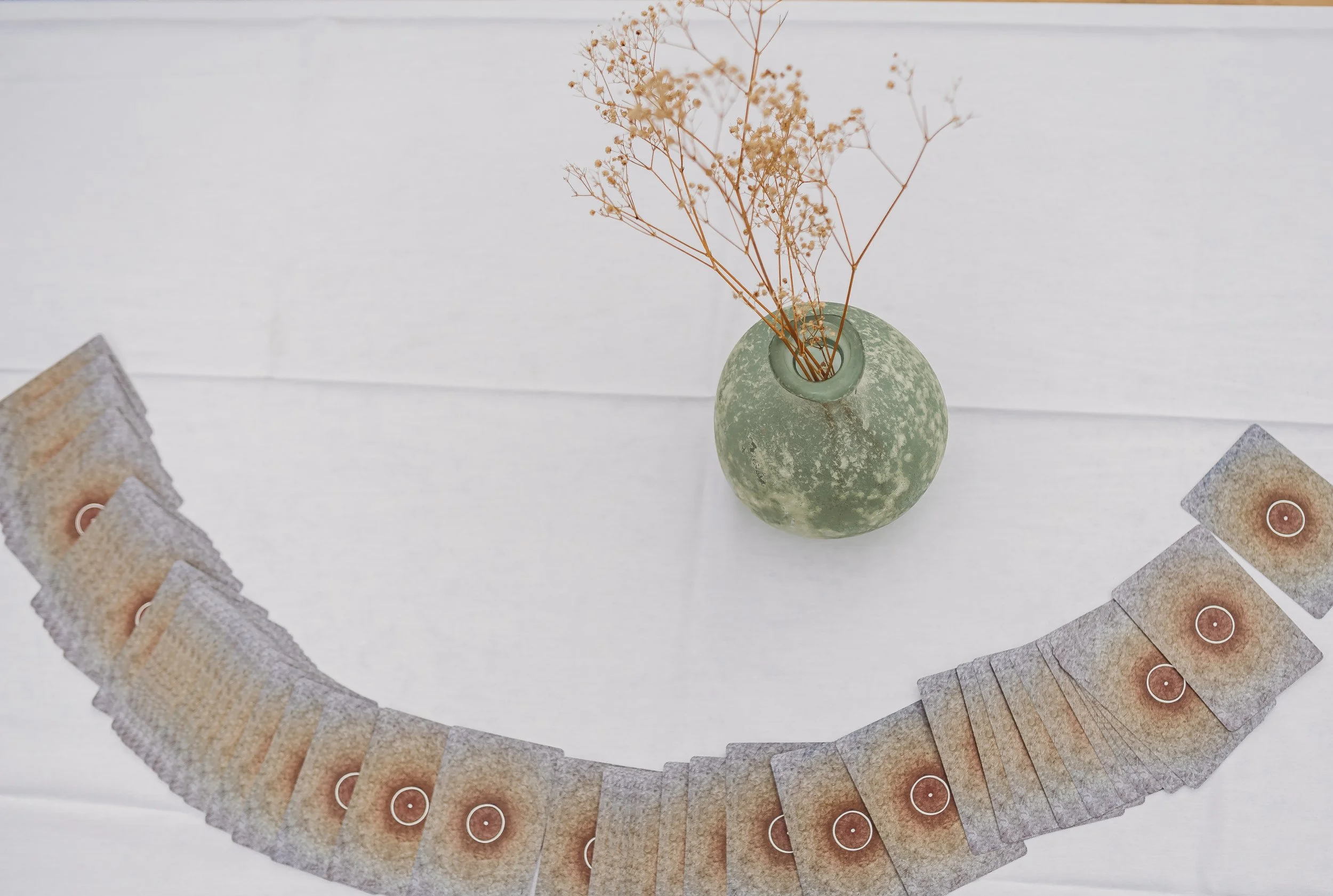A green, textured ceramic vase with a narrow opening, containing dried brown flowers, sits on a white surface. A semi-circle arrangement of playing cards with a red circle in the center surrounds the vase.