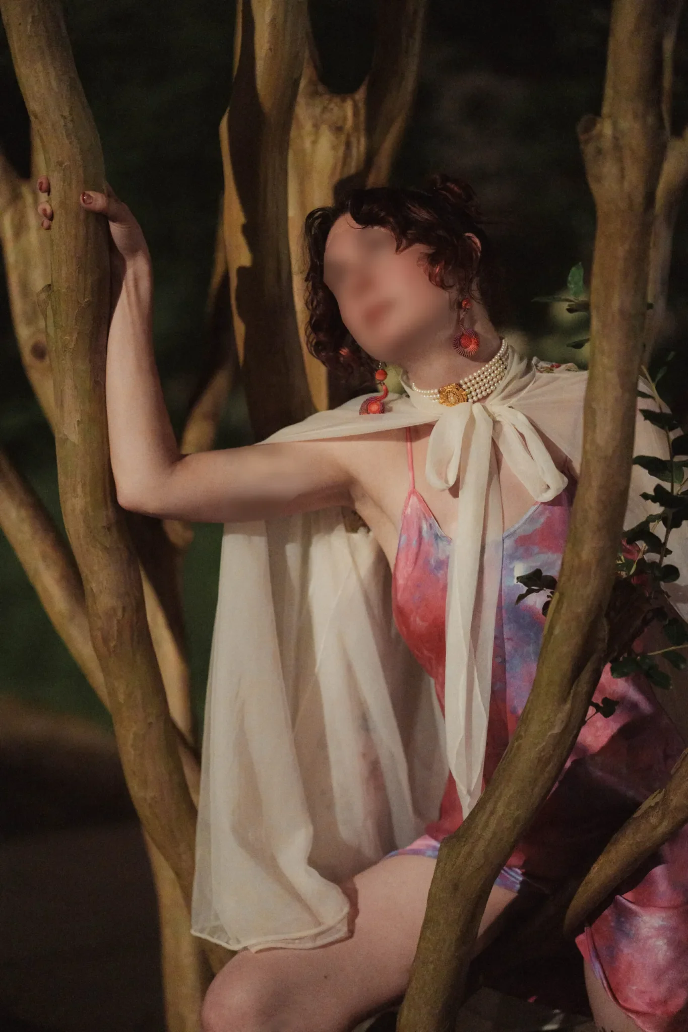 A woman with curly hair, wearing colorful earrings, a pearl necklace, and a cream top, is standing among tree branches in a dark forest or wooded area.