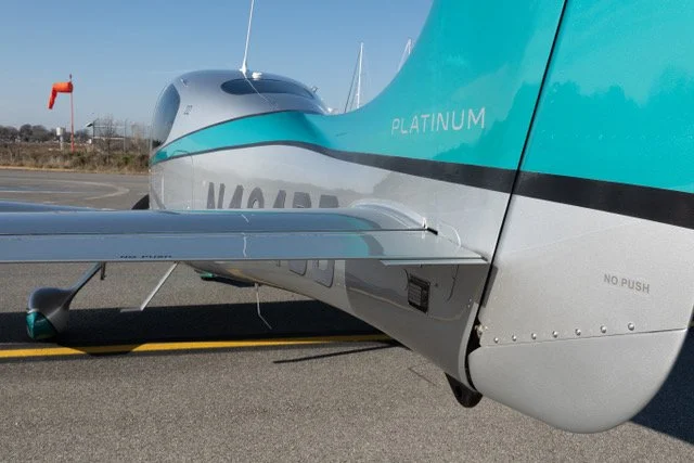Small turquoise and silver airplane on a runway, with "PLATINUM" label on the tail, wing visible, orange windsock in the background.