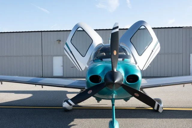 Small teal aircraft with open doors parked in front of a hangar, viewed from the front.