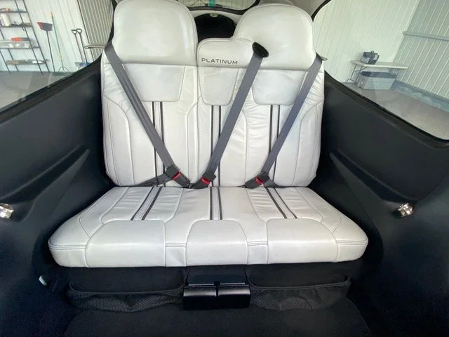 Interior view of a light airplane showing a rear bench seat with white leather upholstery and safety harnesses.