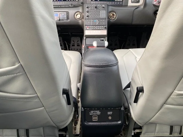 Interior view of an airplane cockpit showing seats, a center console with USB power and audio input, and various control instruments.