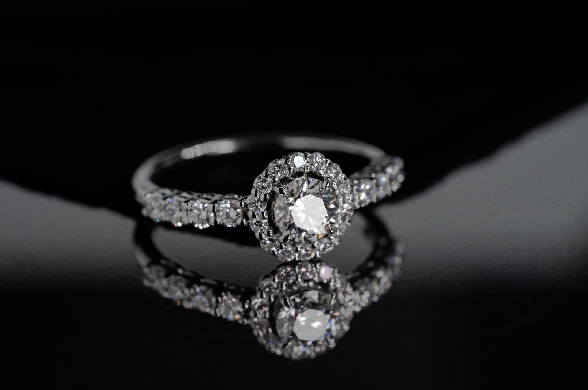 Anillo de compromiso en oro blanco con diamante central de corte oval de 1.00 ct con certificado gemológico, halo de diamantes de corte brillante de 0.14 ct y diamantes laterales de corte brillante de 0.24 ct.