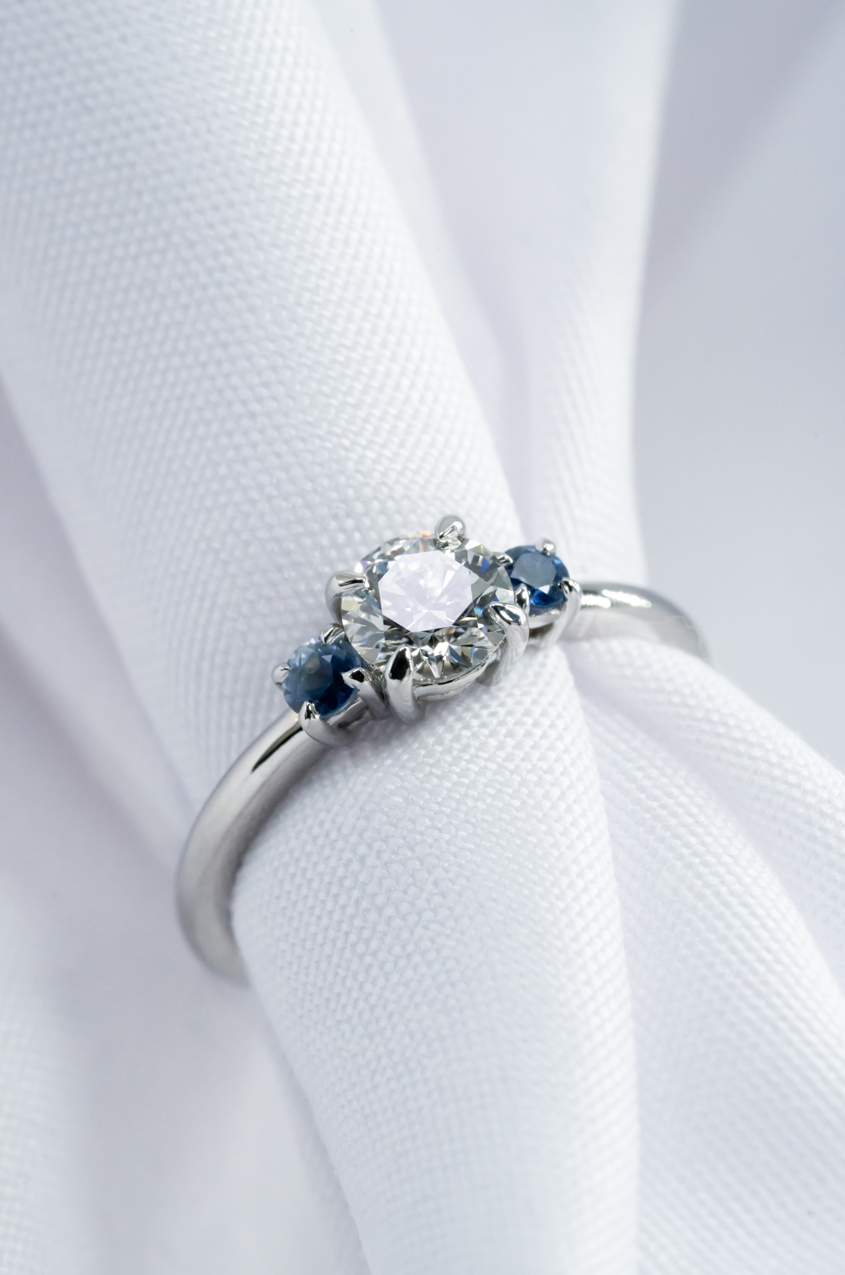 Anillo trilogía en oro blanco con diamante natural central de corte brillante y zafiros naturales laterales de corte redondo.