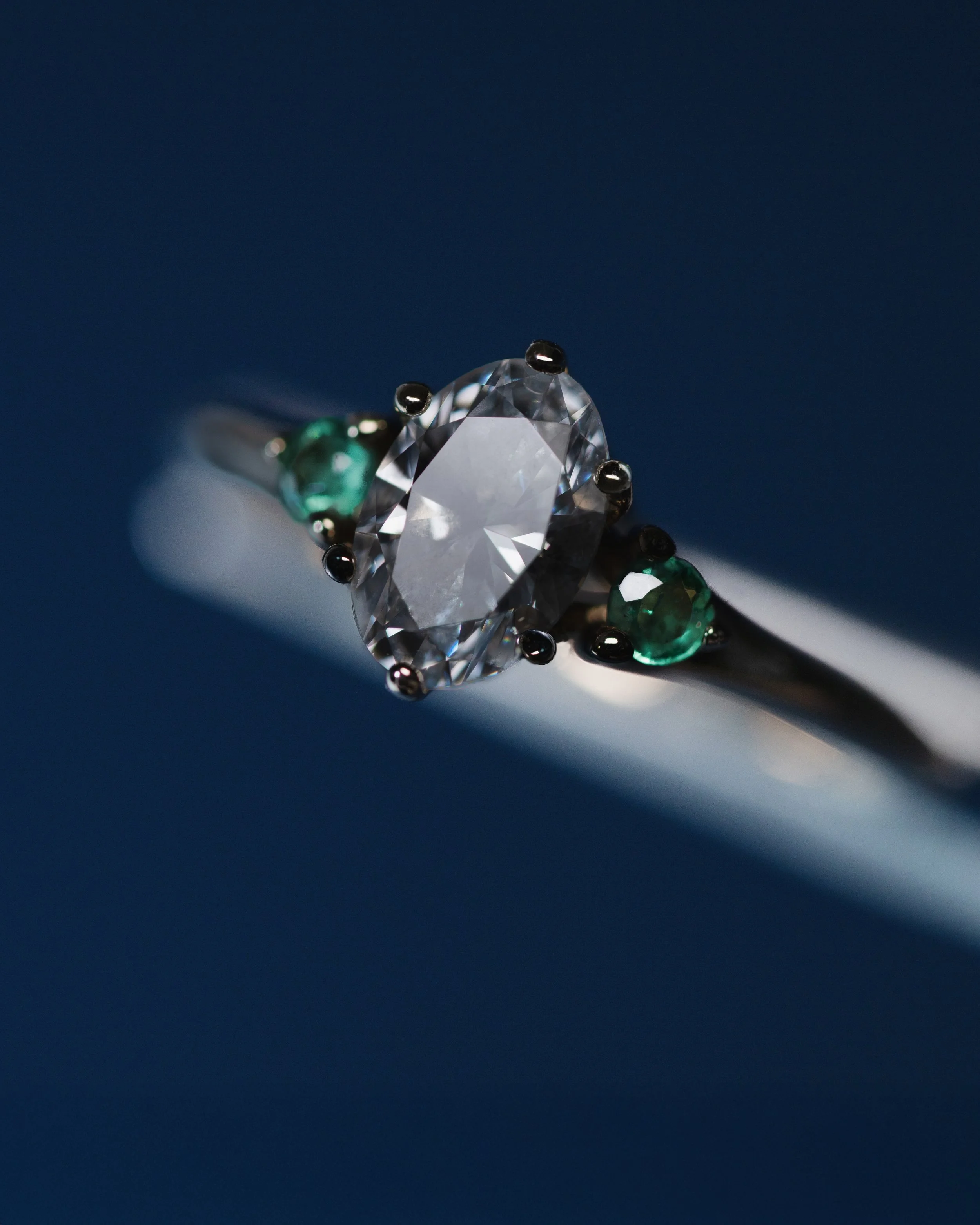 Anillo trilogía en oro blanco con diamante central de corte oval de 1.00 ct con certificado gemológico y esmeraldas naturales laterales de corte redondo.
