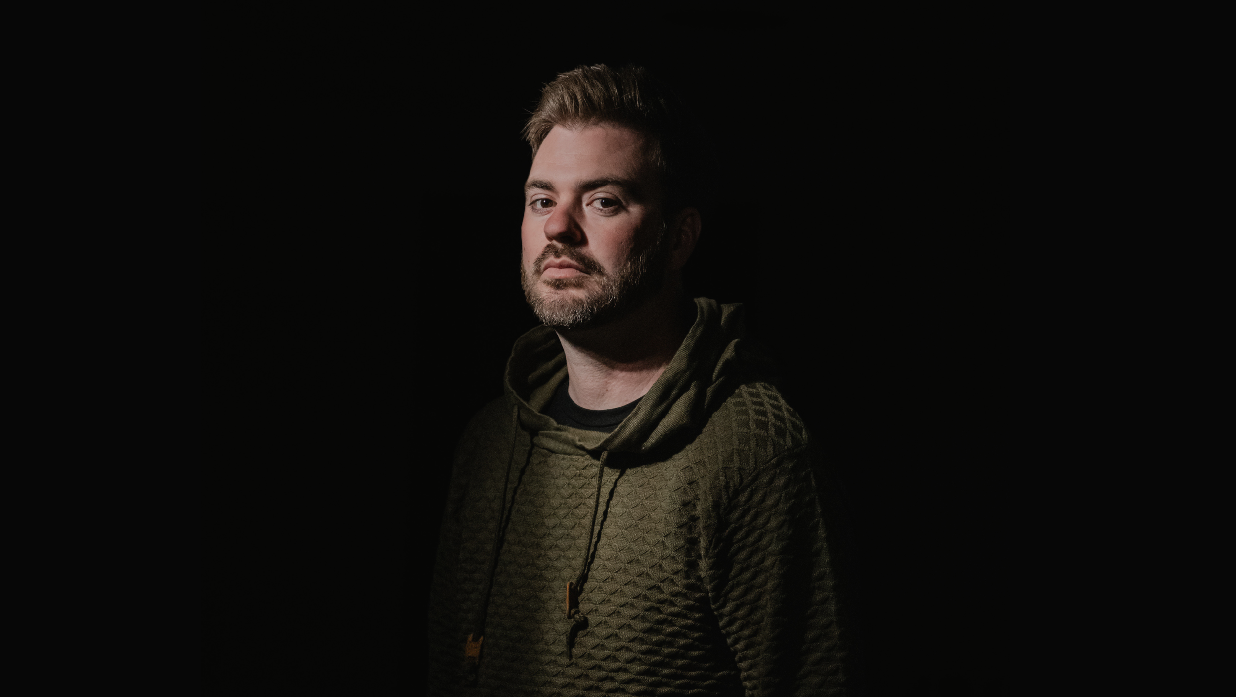 Portrait of a man with short hair and a beard, wearing a patterned hoodie, against a black background.