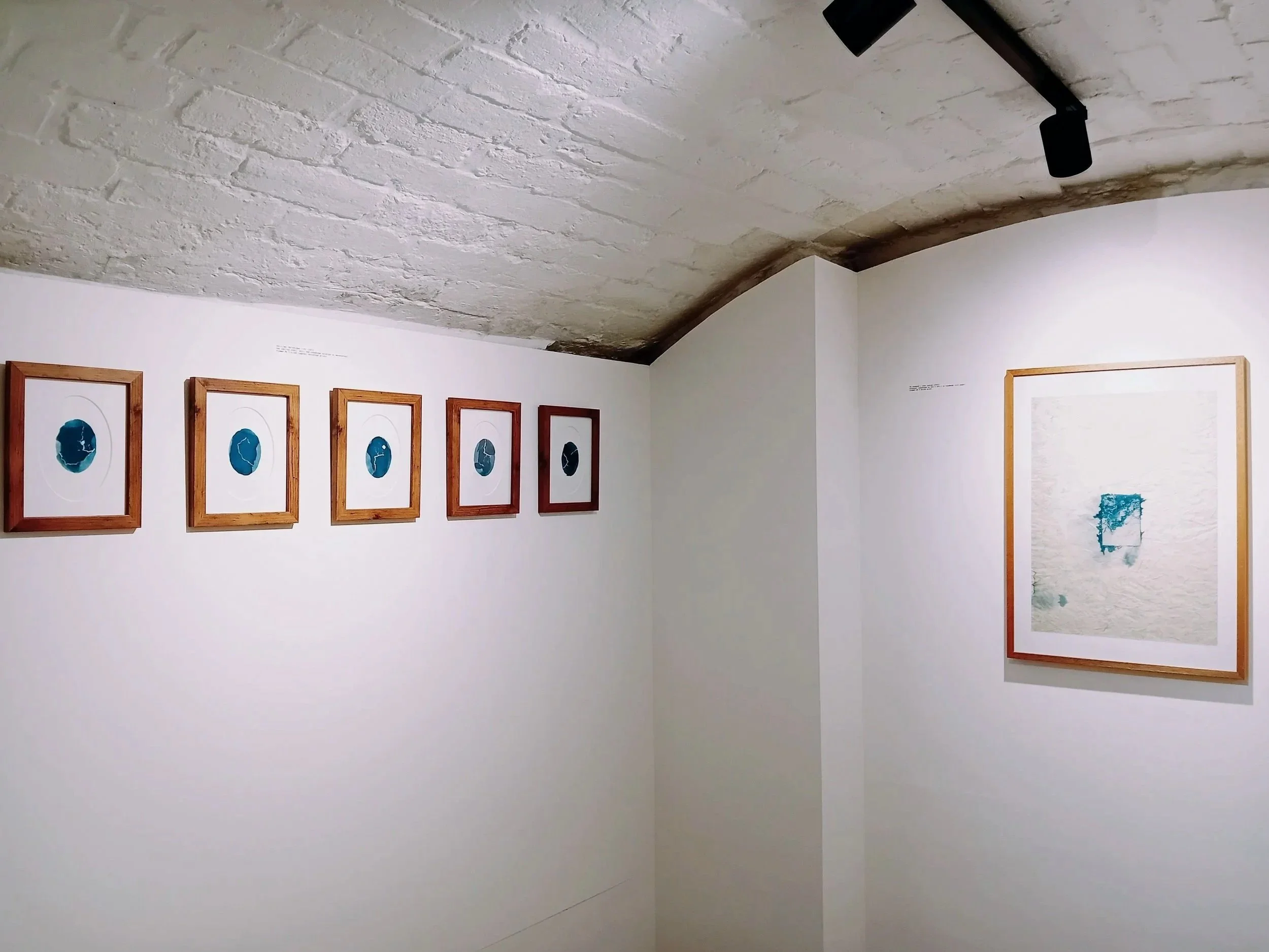 Gallery wall with seven framed blue abstract artworks inside an art gallery, white brick ceiling, and white walls.