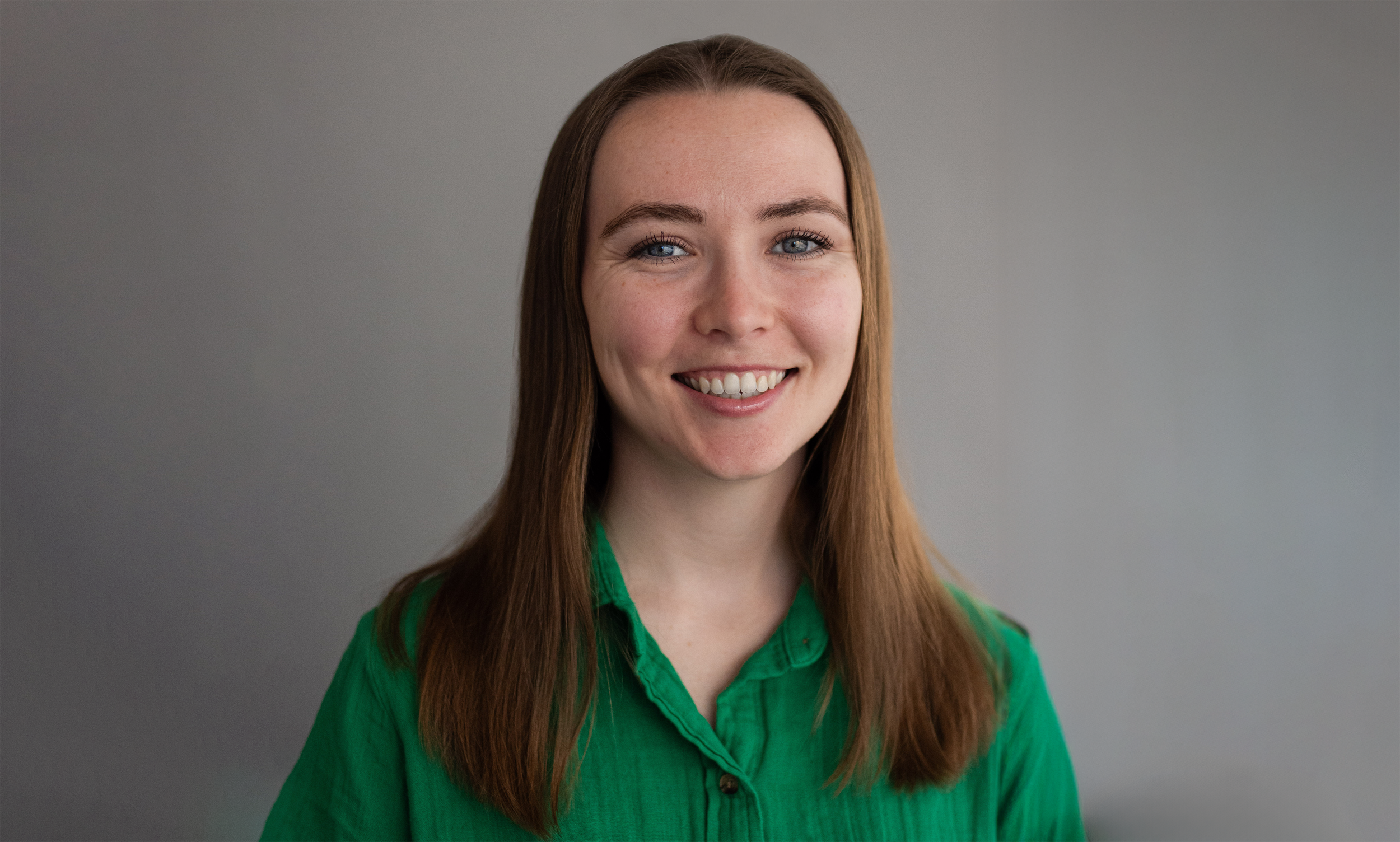 Portrait of a young woman with long brown hair, wearing a green shirt, smiling against a plain gray background.