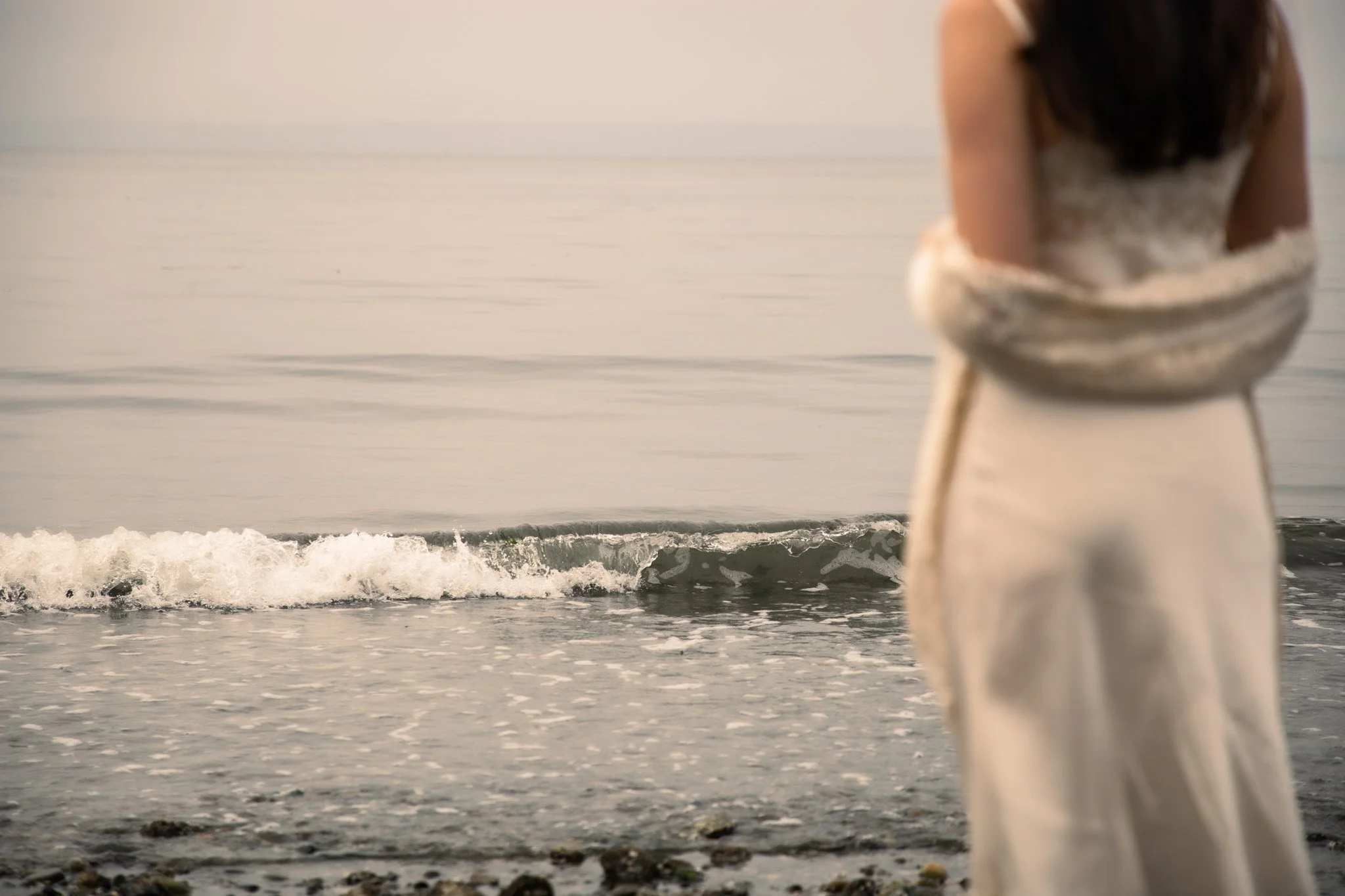 Person sitting on the beach near the water, with ocean and waves in the background, seen from behind.