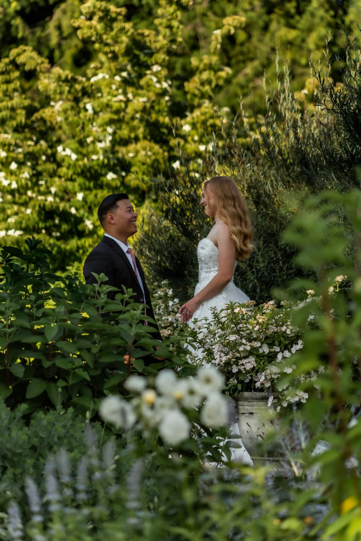 A bride and groom standing outdoors among green bushes and white flowers, gazing at each other during their wedding ceremony.