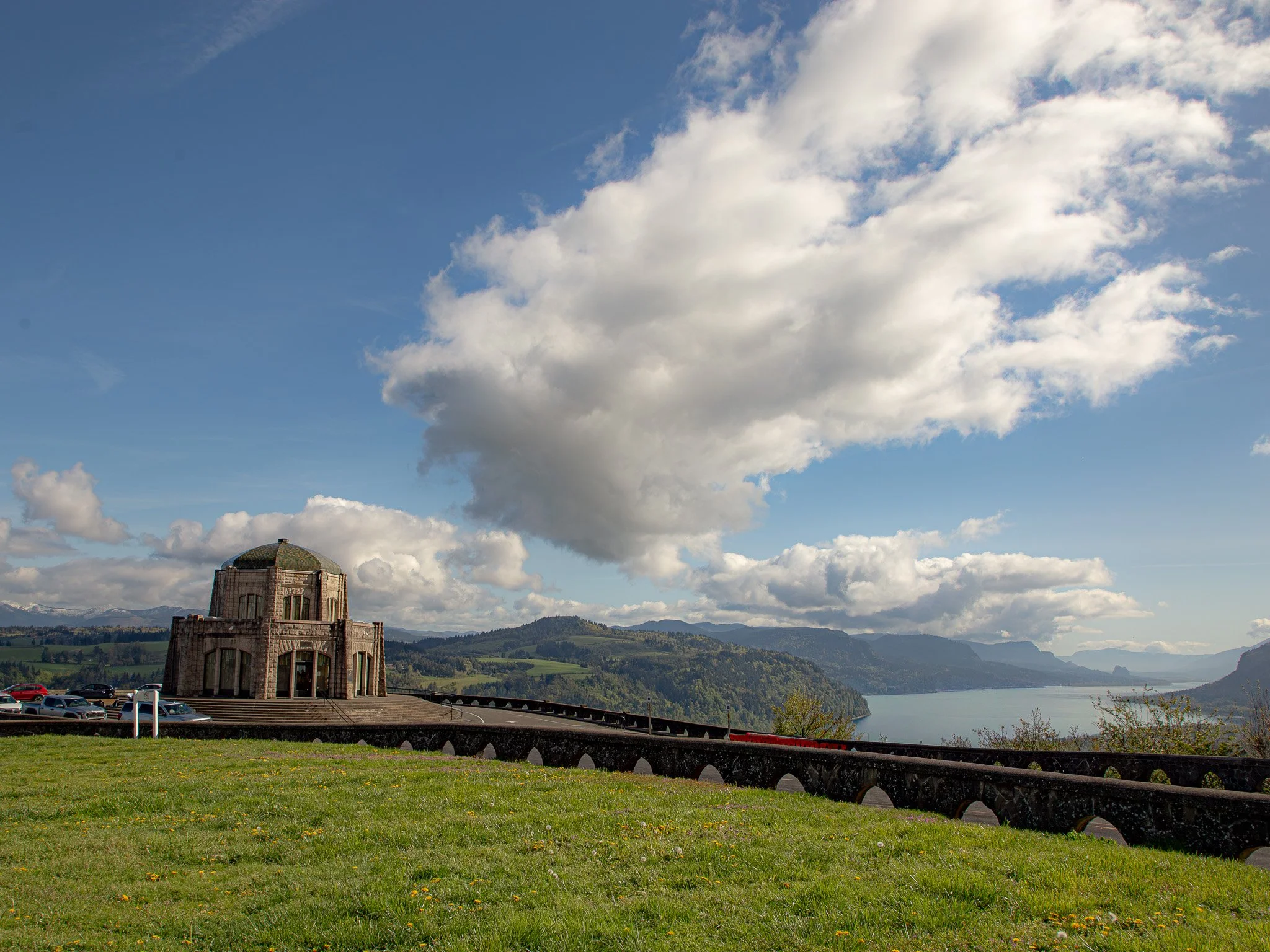 A scenic view of a large body of water with a mountainous landscape in the background under a mostly cloudy sky. In the foreground, there is a green grassy area with yellow flowers, and a stone building with a dome-shaped roof and large windows. A parking lot with cars and an EV charging station is situated near the building.