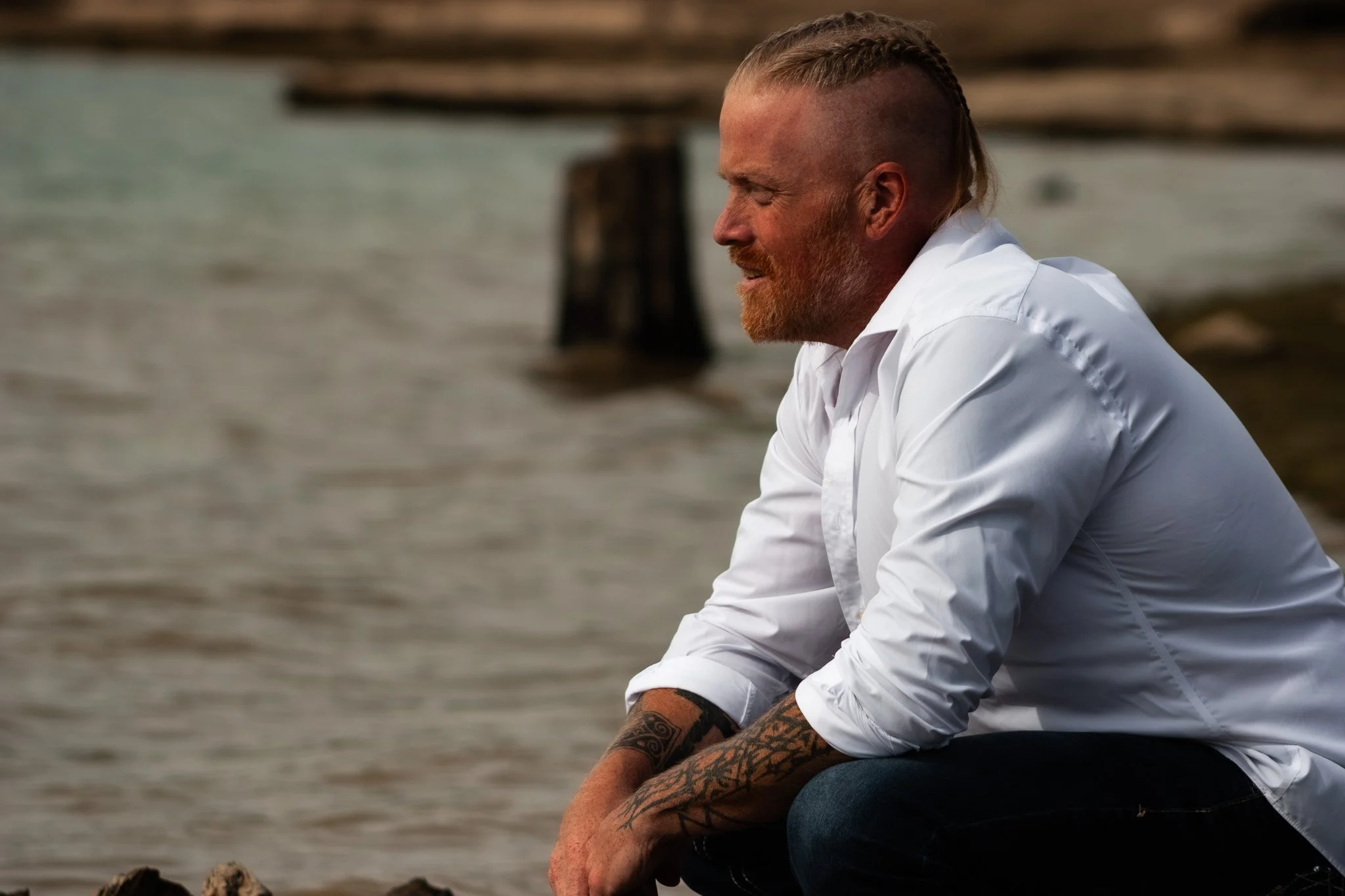 A man with a beard and tattoos wearing a white shirt, sitting by the water with logs or posts in the background, looking to the right with a contemplative expression.