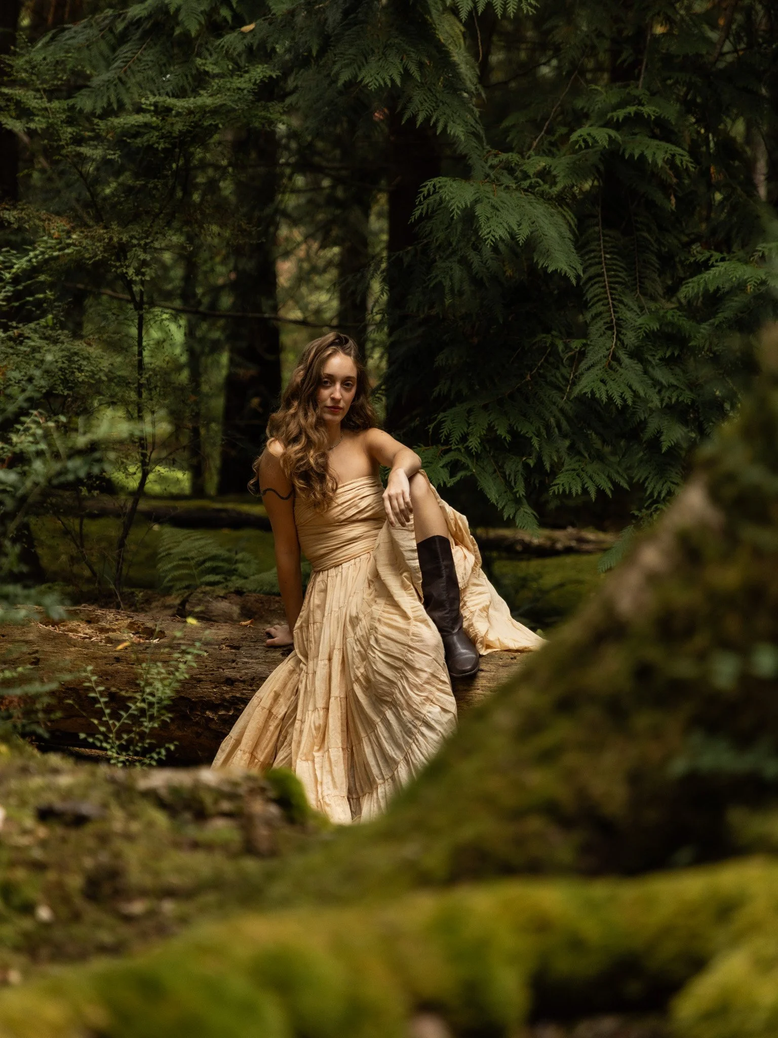 A woman with long, curly brown hair and fair skin, wearing a beige strapless dress and black boots, sitting on a log in a lush green forest.