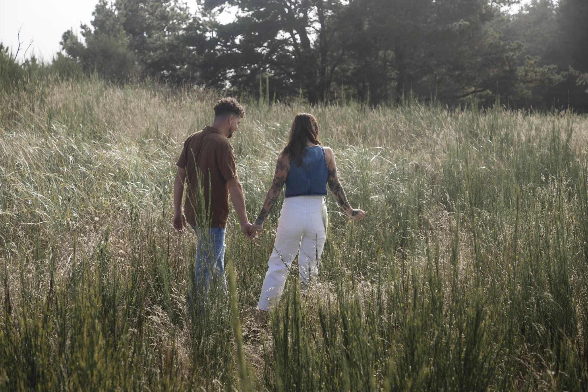 A couple holding hands walking through a grassy field with trees in the background.