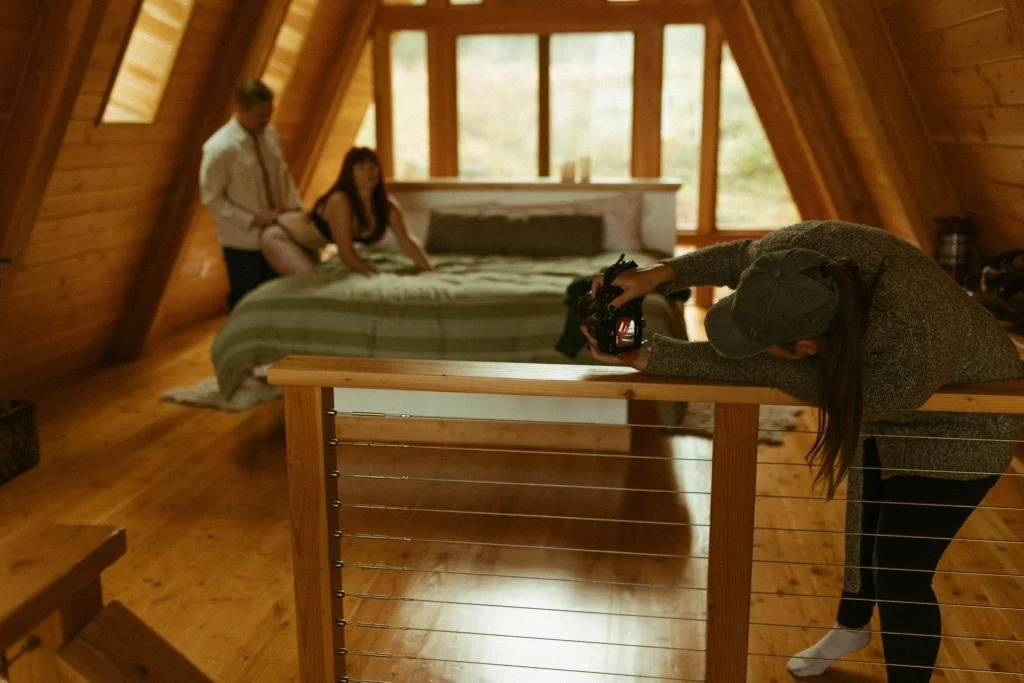 A woman with a cap and long hair taking a photo of a man and woman sitting on a bed in an A-frame cabin interior with wooden walls and floor.