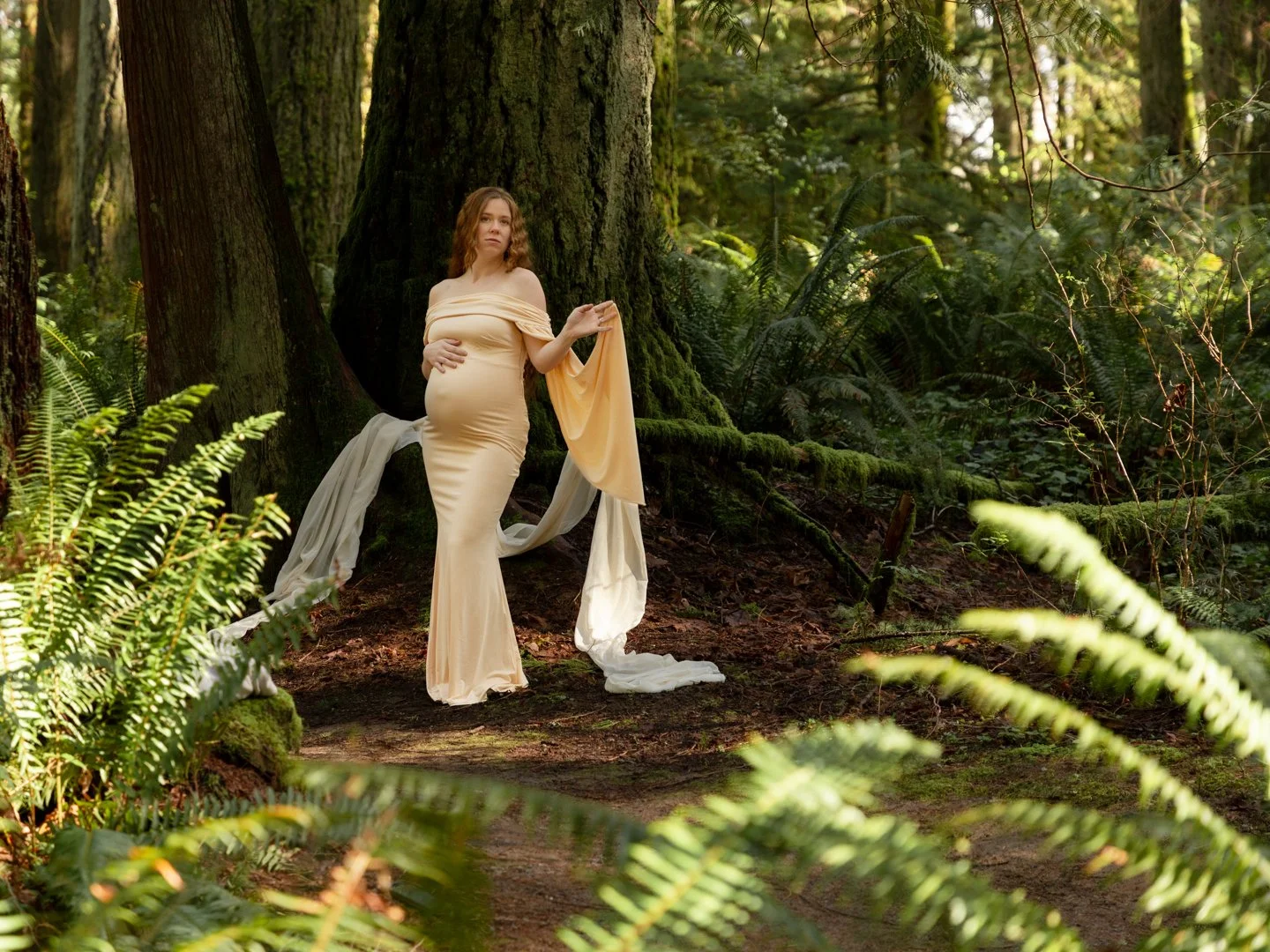 A pregnant woman in an elegant, flowing cream-colored gown stands in a lush forest, leaning against a large moss-covered tree with ferns and greenery surrounding her.