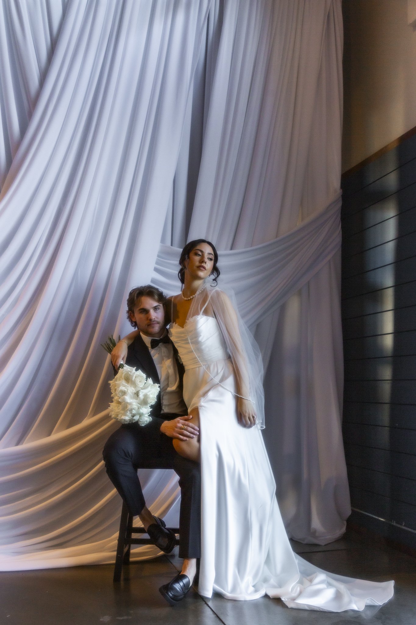 A bride in a white wedding dress standing next to a groom in a black suit, with a sheer veil and holding a white bouquet, posing in front of white draped fabric backdrop at a wedding or formal event.