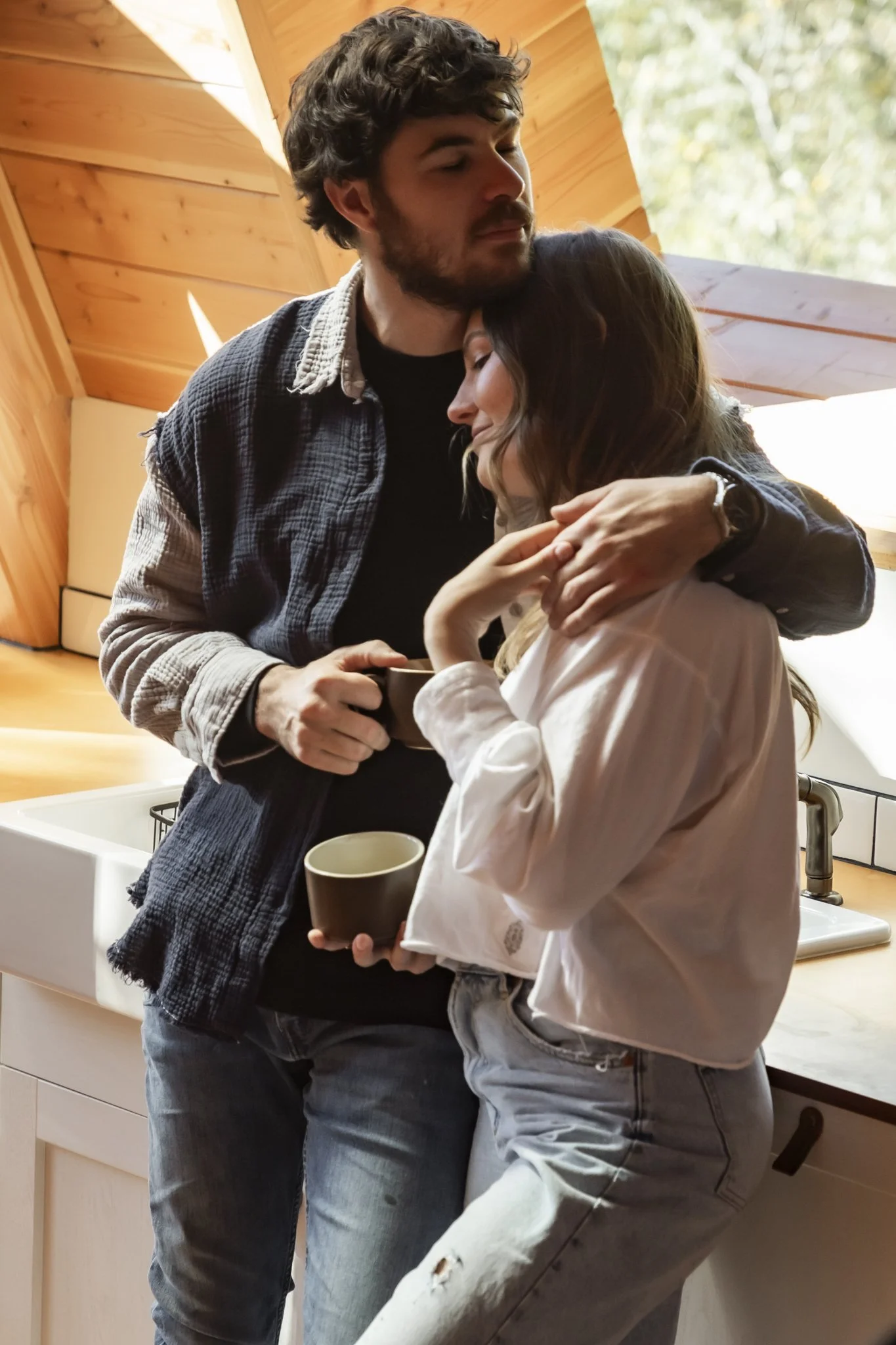 A man and woman hugging in a kitchen, holding coffee mugs, with wooden ceiling and windows with trees outside.