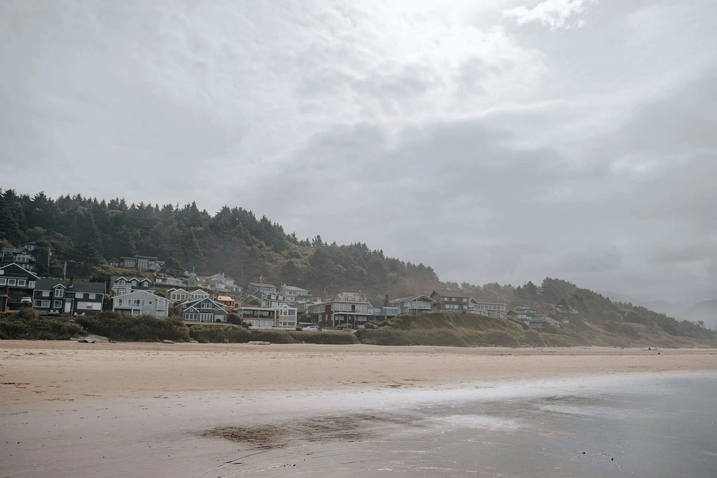 Cloudy sky over a coastal neighborhood with houses on a hillside and a sandy beach in the foreground.