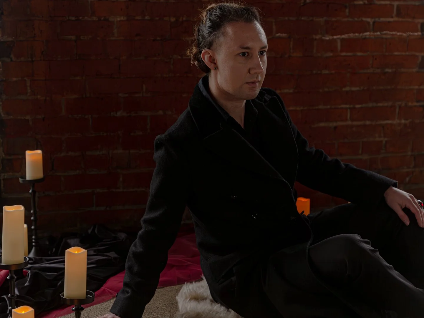 A person with curly hair sitting on the floor near a brick wall, surrounded by candles, wearing a black jacket and black pants.