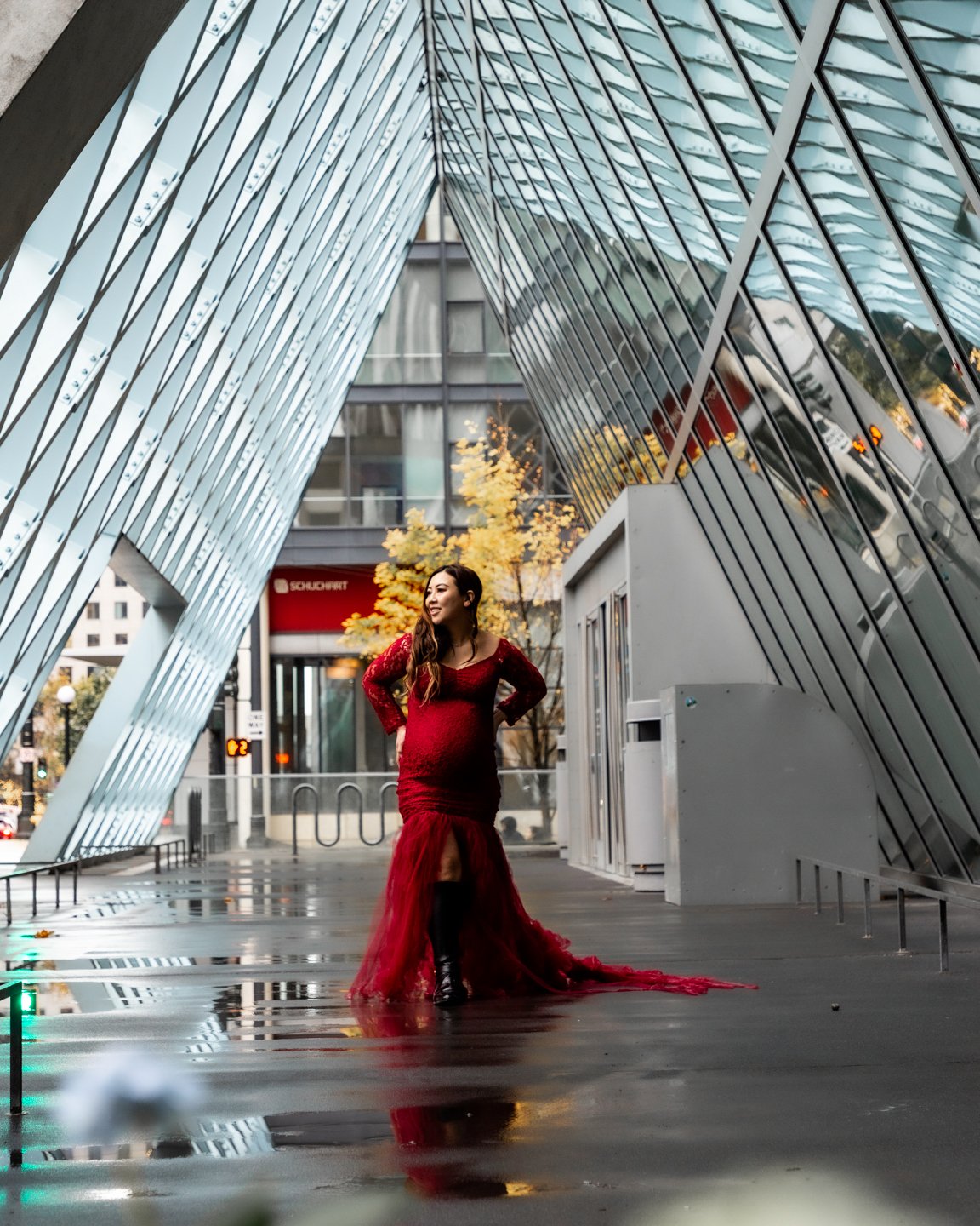 A woman in a red gown standing outdoors on a city street with modern architecture and trees in the background.