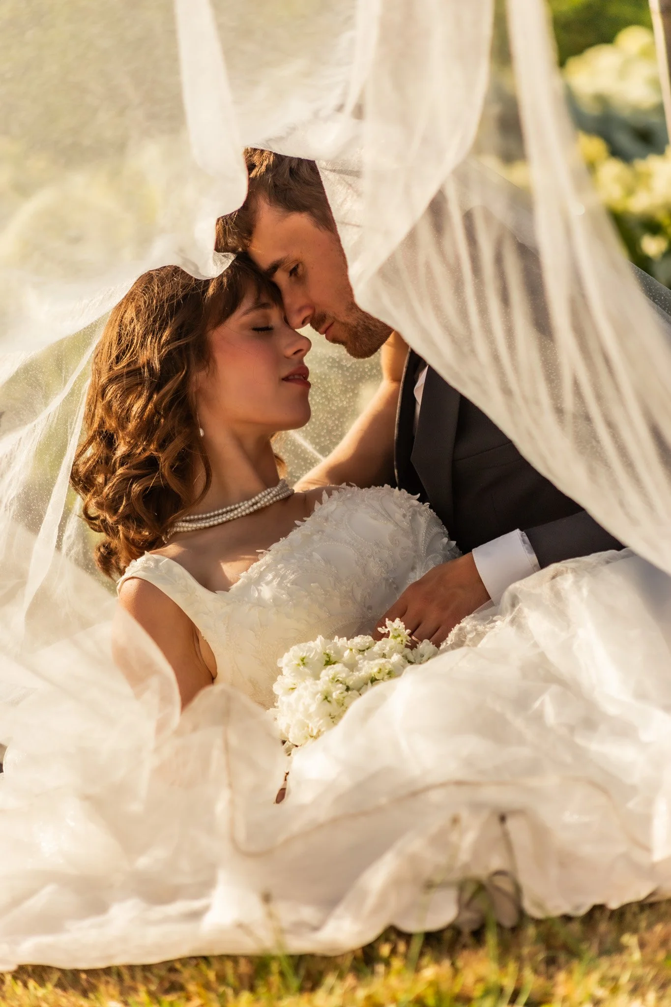 A bride and groom are seen with their foreheads touching, eyes closed, under a flowing white veil, holding a bouquet of white flowers, in an outdoor setting.